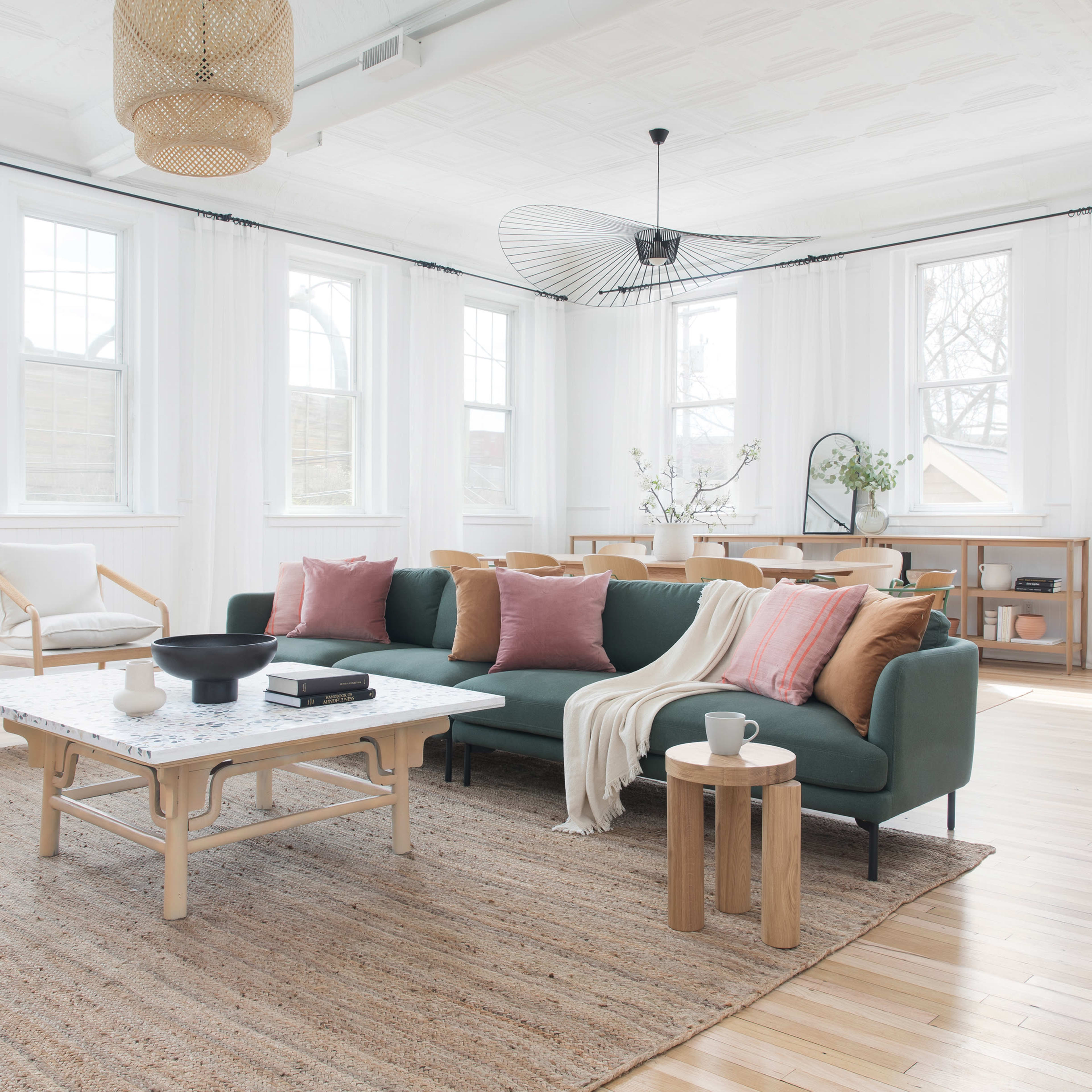 A spacious living room features a green sofa with pillows, a white coffee table, natural wood furnishings, and large windows letting in ample light.