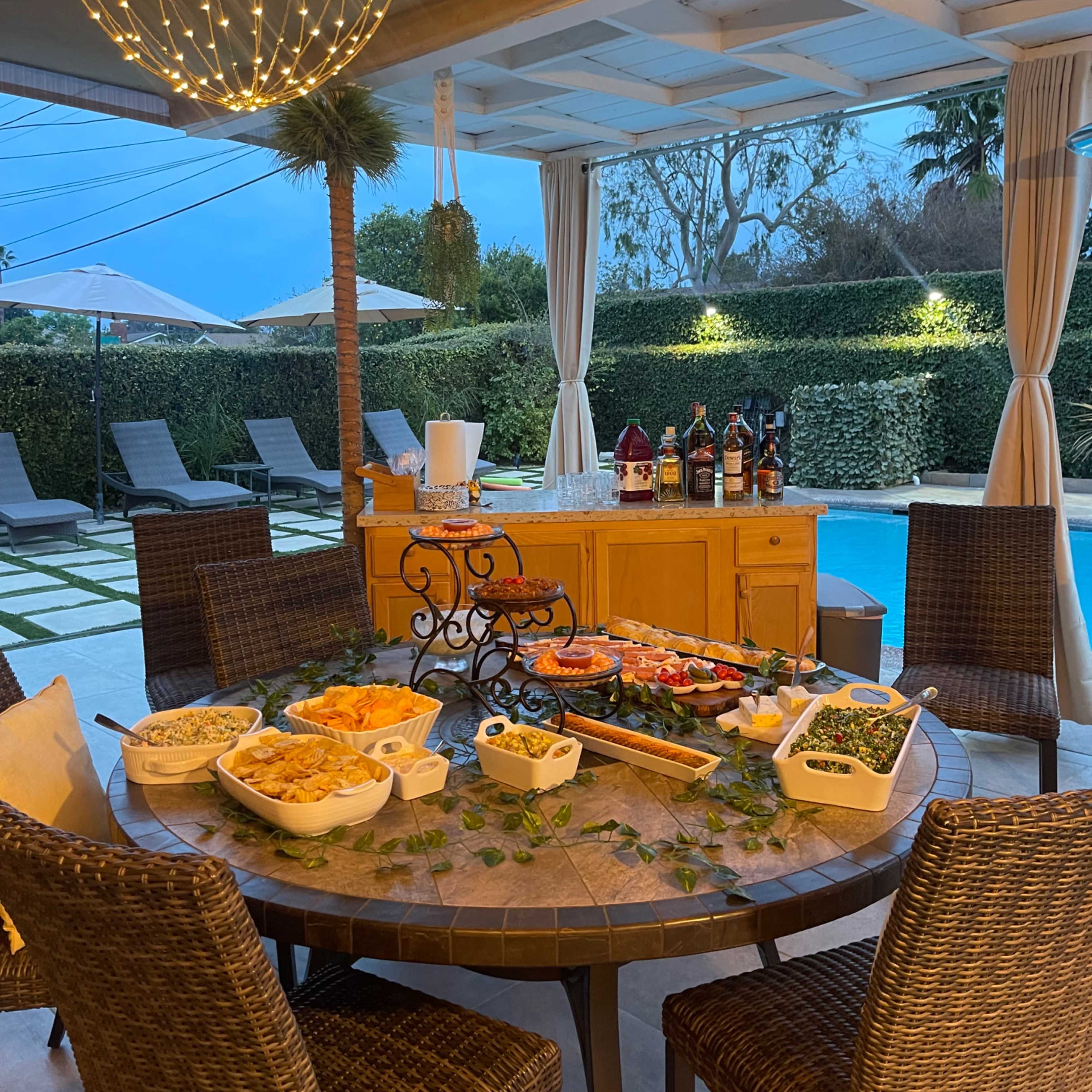 A table with various dishes is set up beneath a covered patio, next to a swimming pool surrounded by lounge chairs.