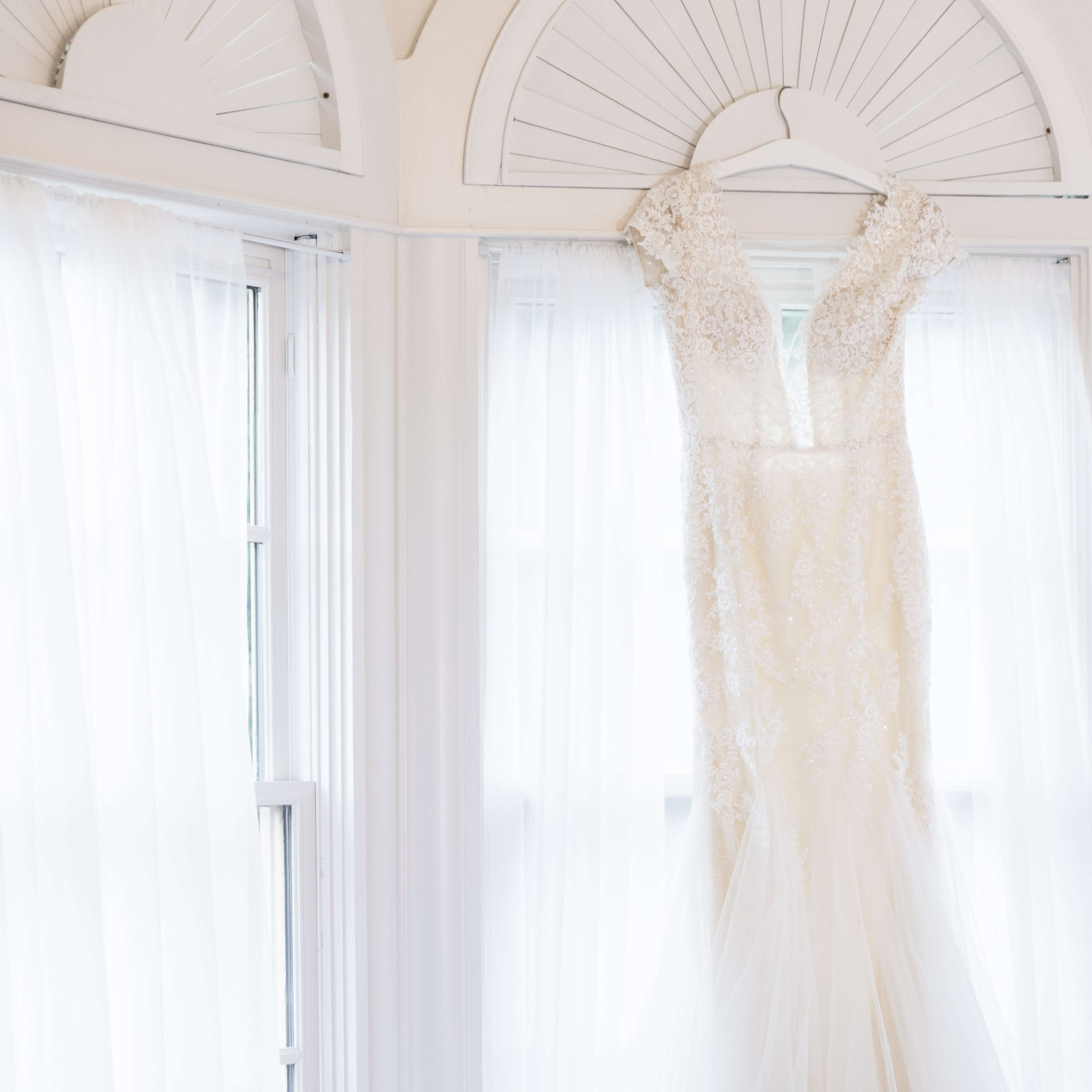 A bridal gown hangs on a hanger against a backdrop of flowing white curtains.