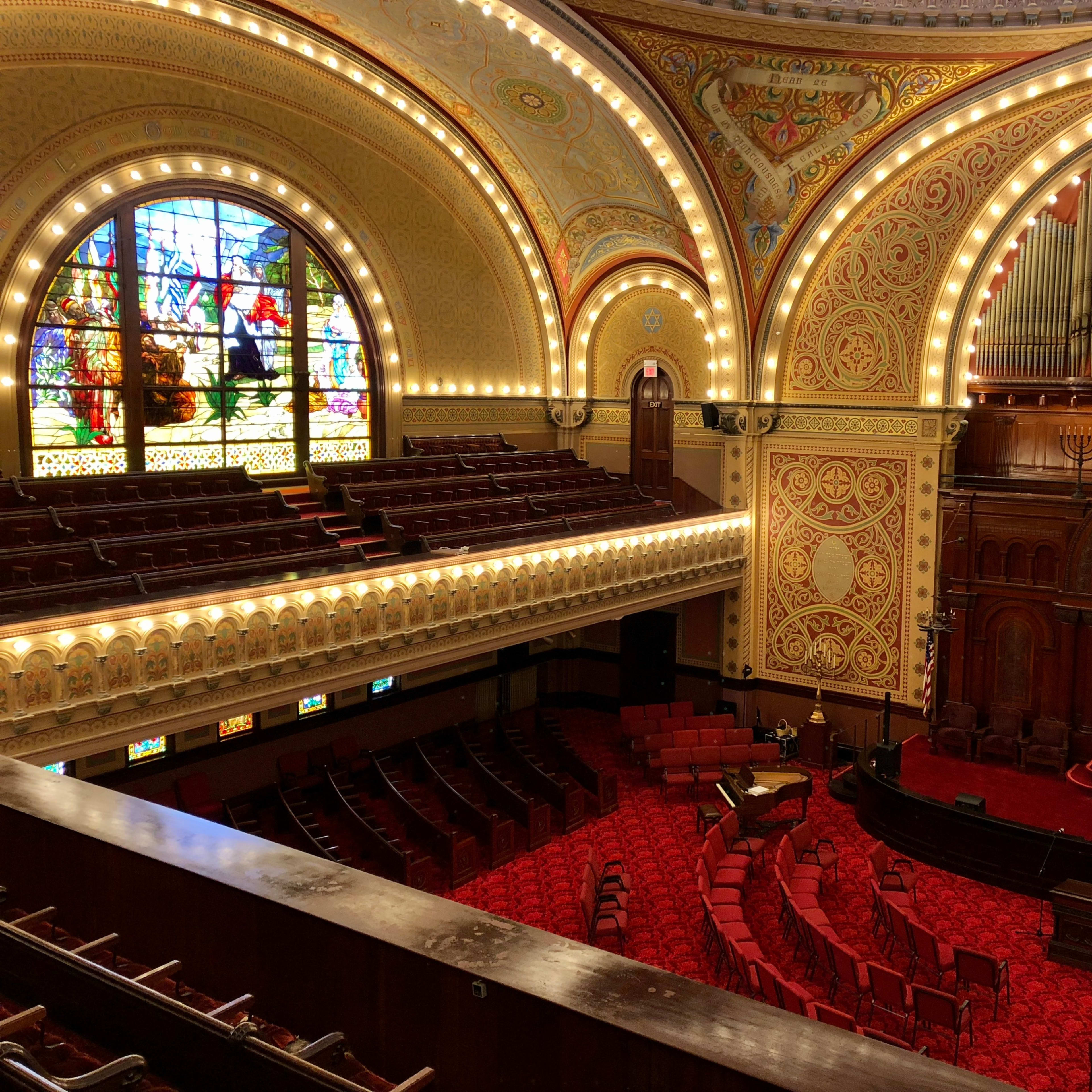 An ornate legislative chamber with red seating, a decorative ceiling, and a large stained glass window.