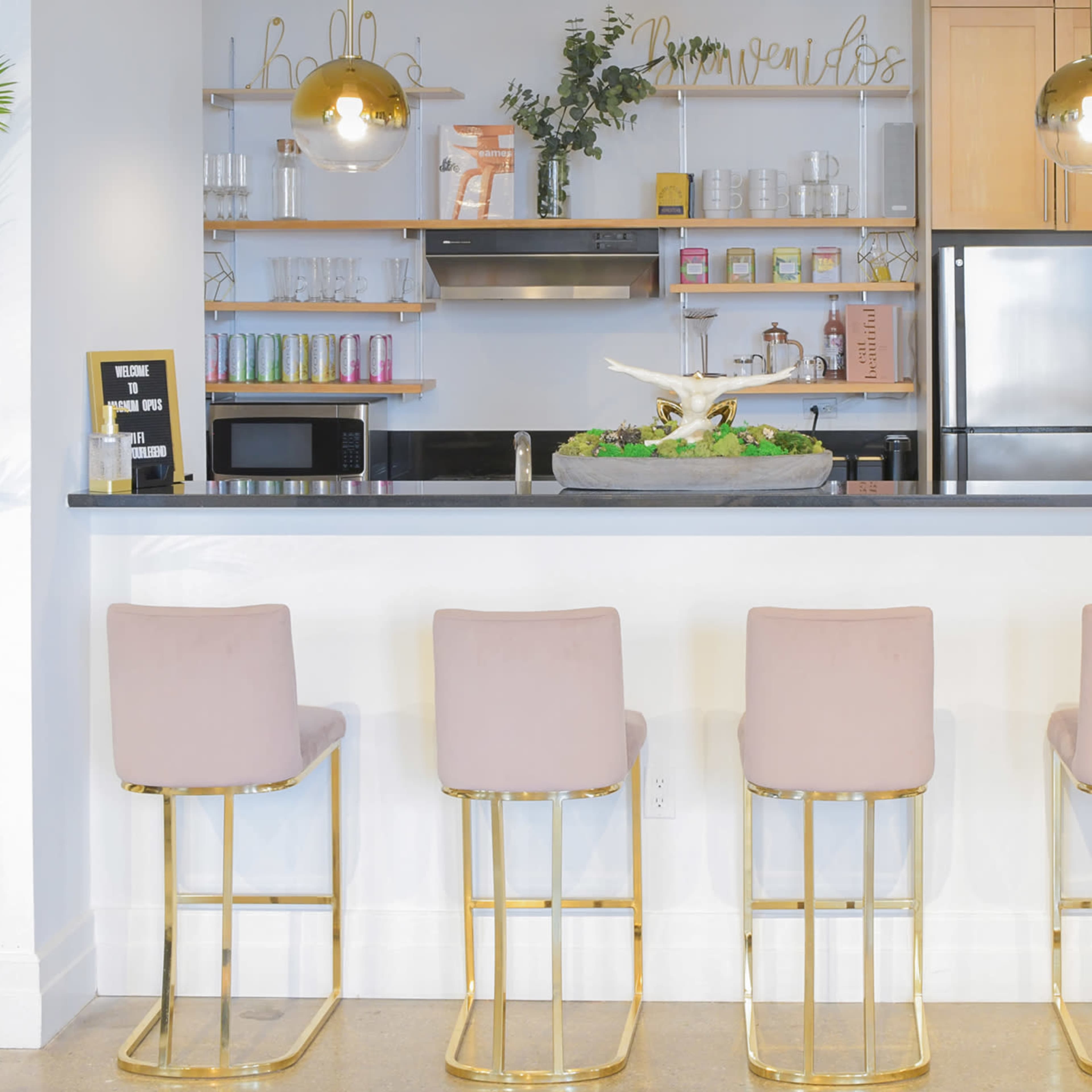 A modern kitchen bar area with four pink upholstered stools and a countertop featuring a variety of kitchen appliances and decorative items.