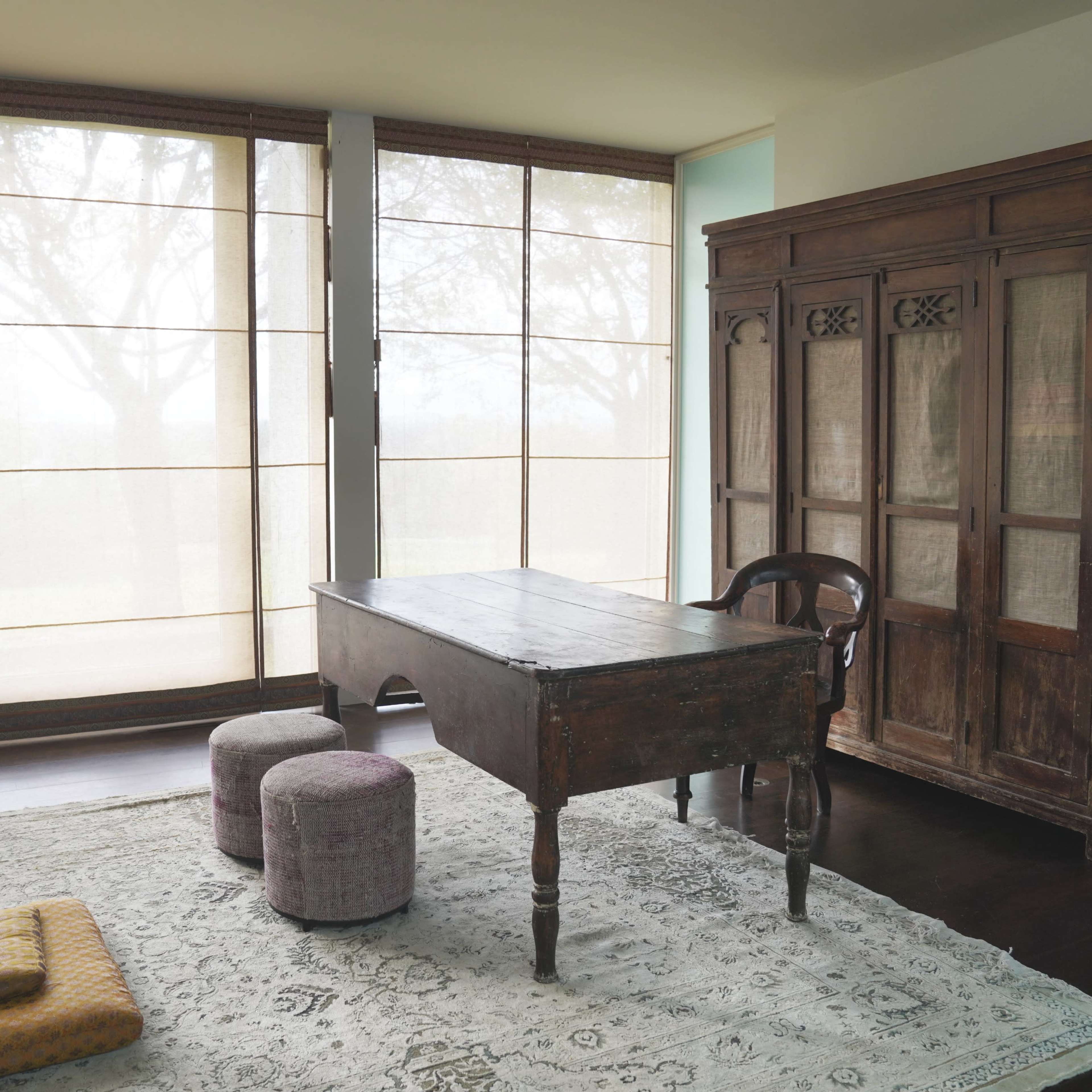 A wooden desk sits in a well-lit room with large windows and a vintage cabinet, accompanied by soft seating on a patterned rug.