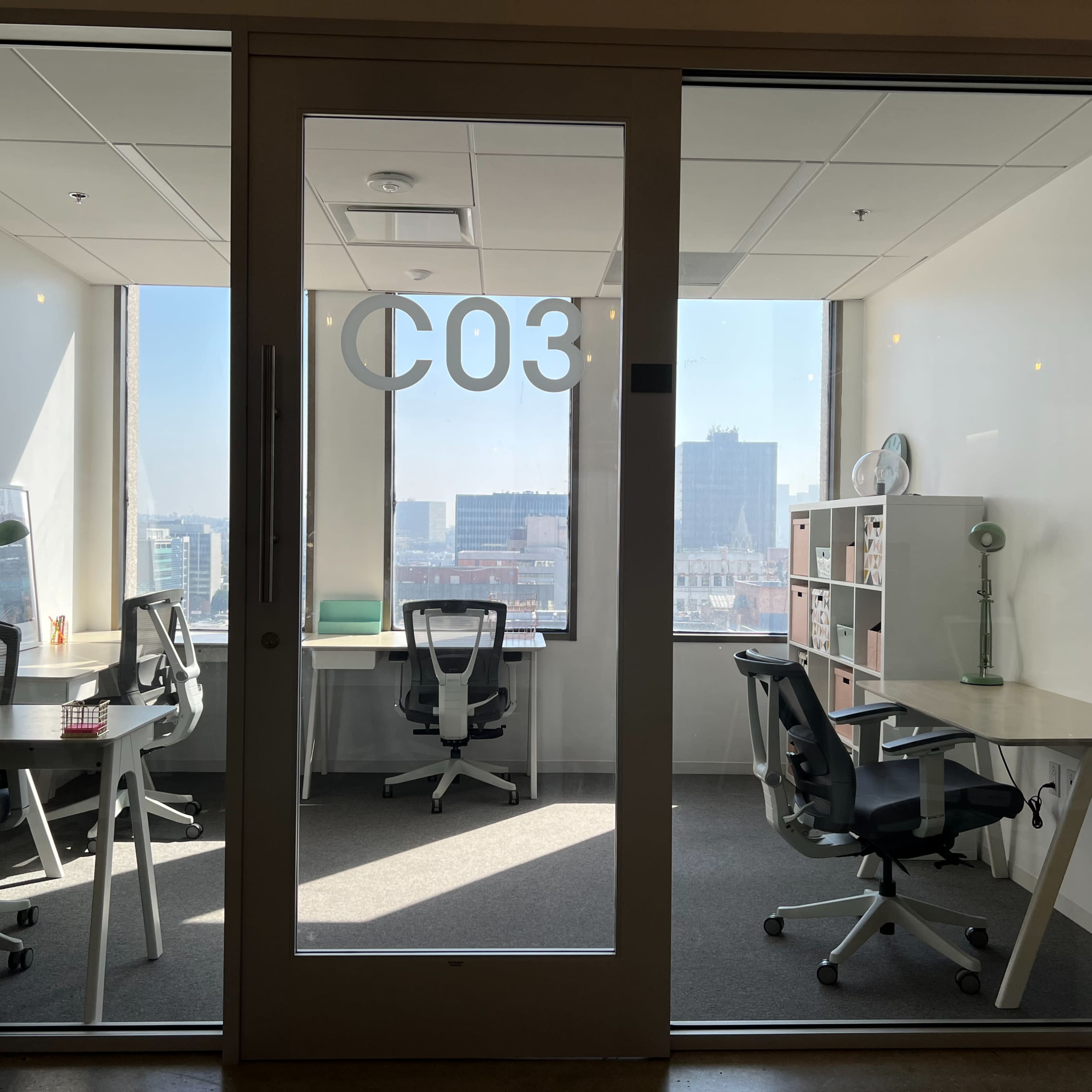 A glass-walled office space labeled "C03" with two desks, each equipped with a chair and a view of the city skyline outside.