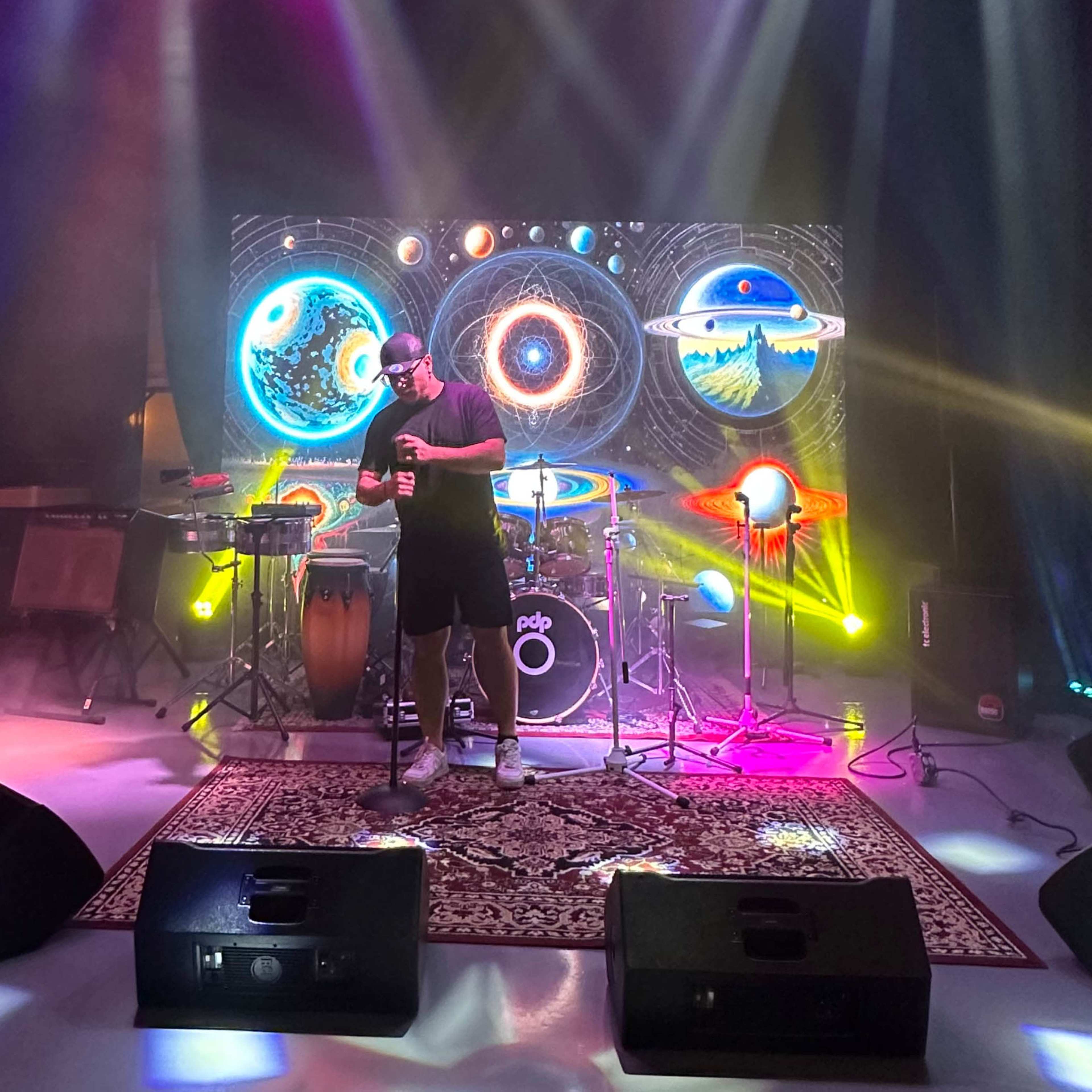A musician performs on stage in front of a colorful cosmic backdrop, accompanied by various instruments and stage lights.
