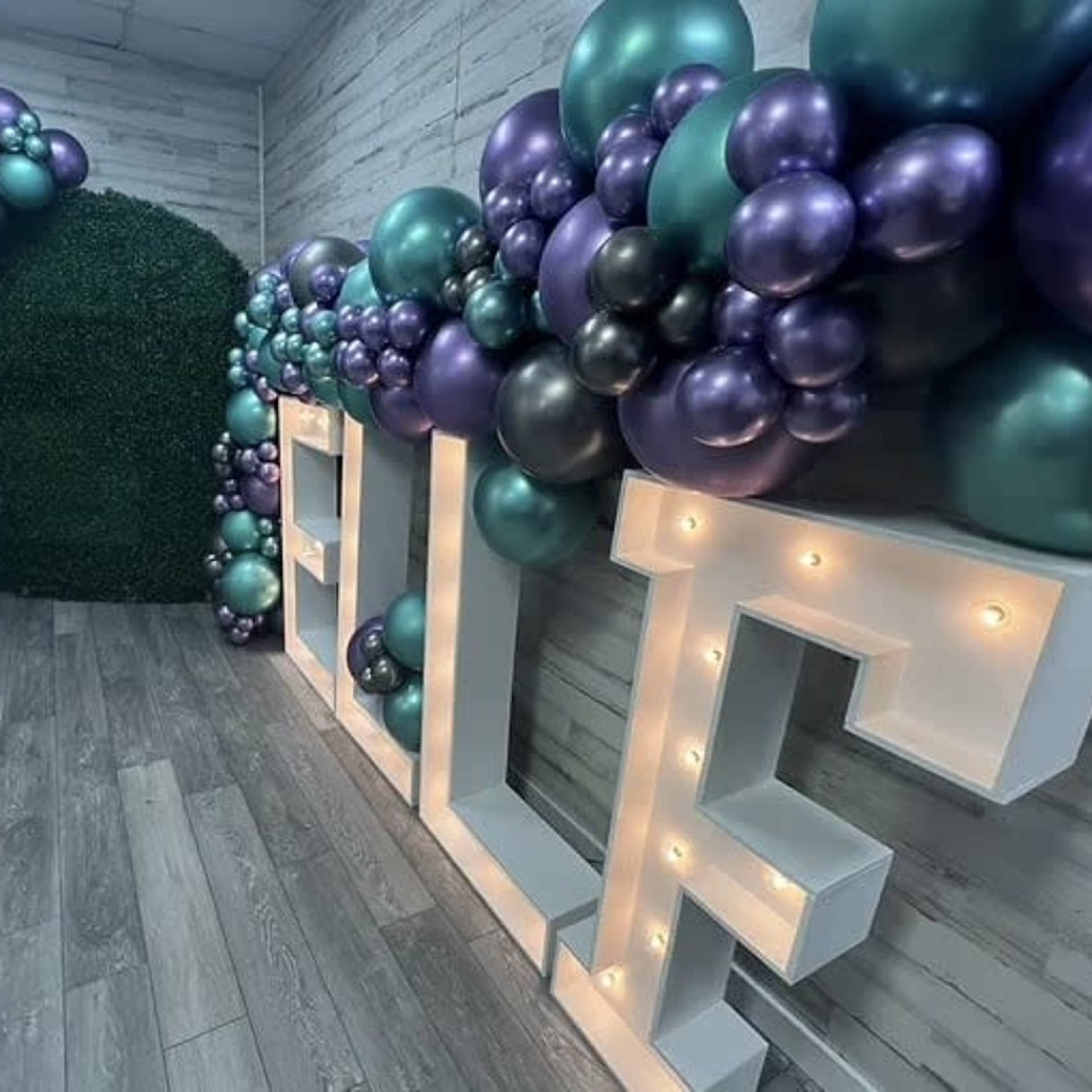 A decorated space features large illuminated letters spelling "SELF" alongside clusters of purple and teal balloons, paired with a green floral backdrop.