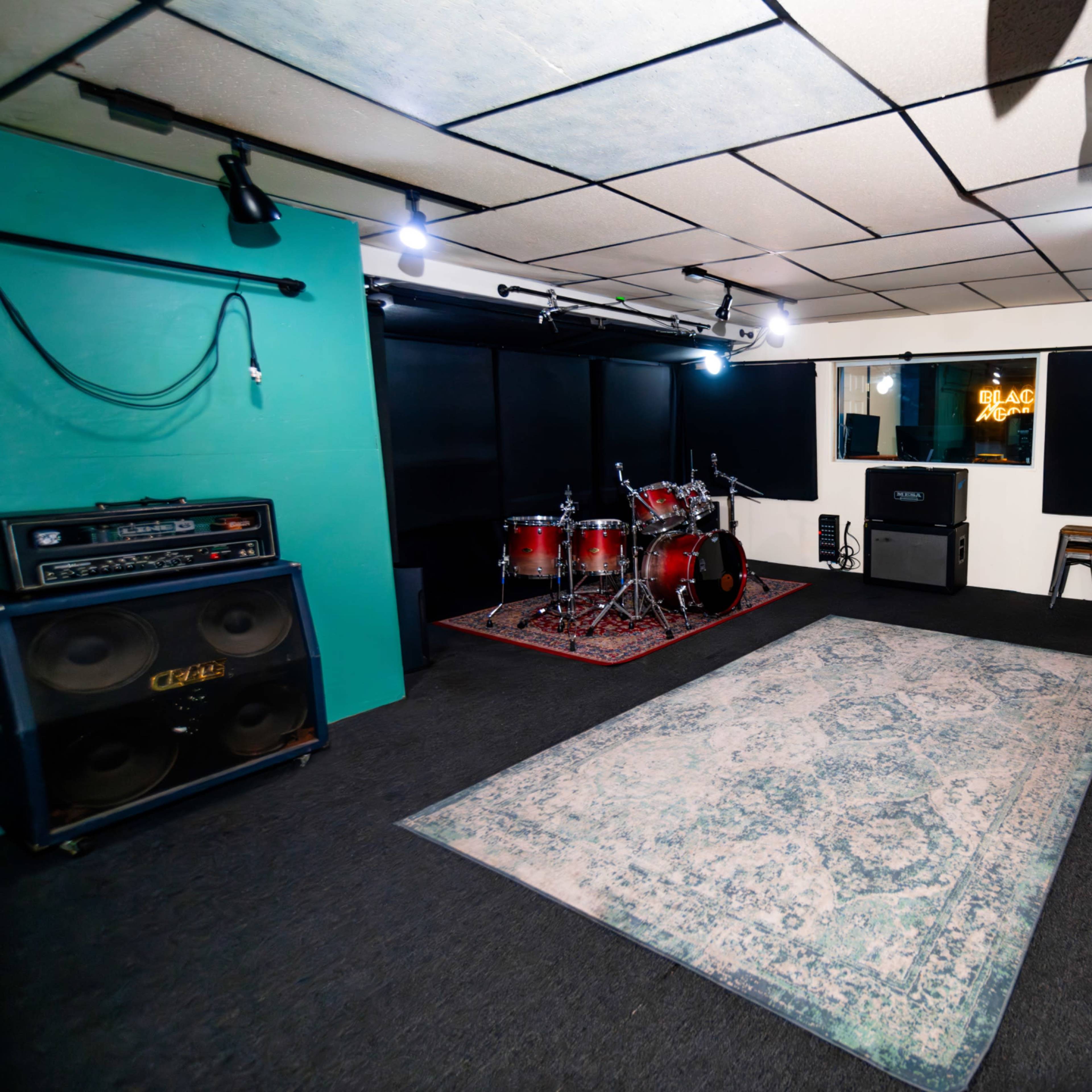 A music rehearsal studio with a drum set, amplifiers, and sound equipment arranged on a carpeted floor.