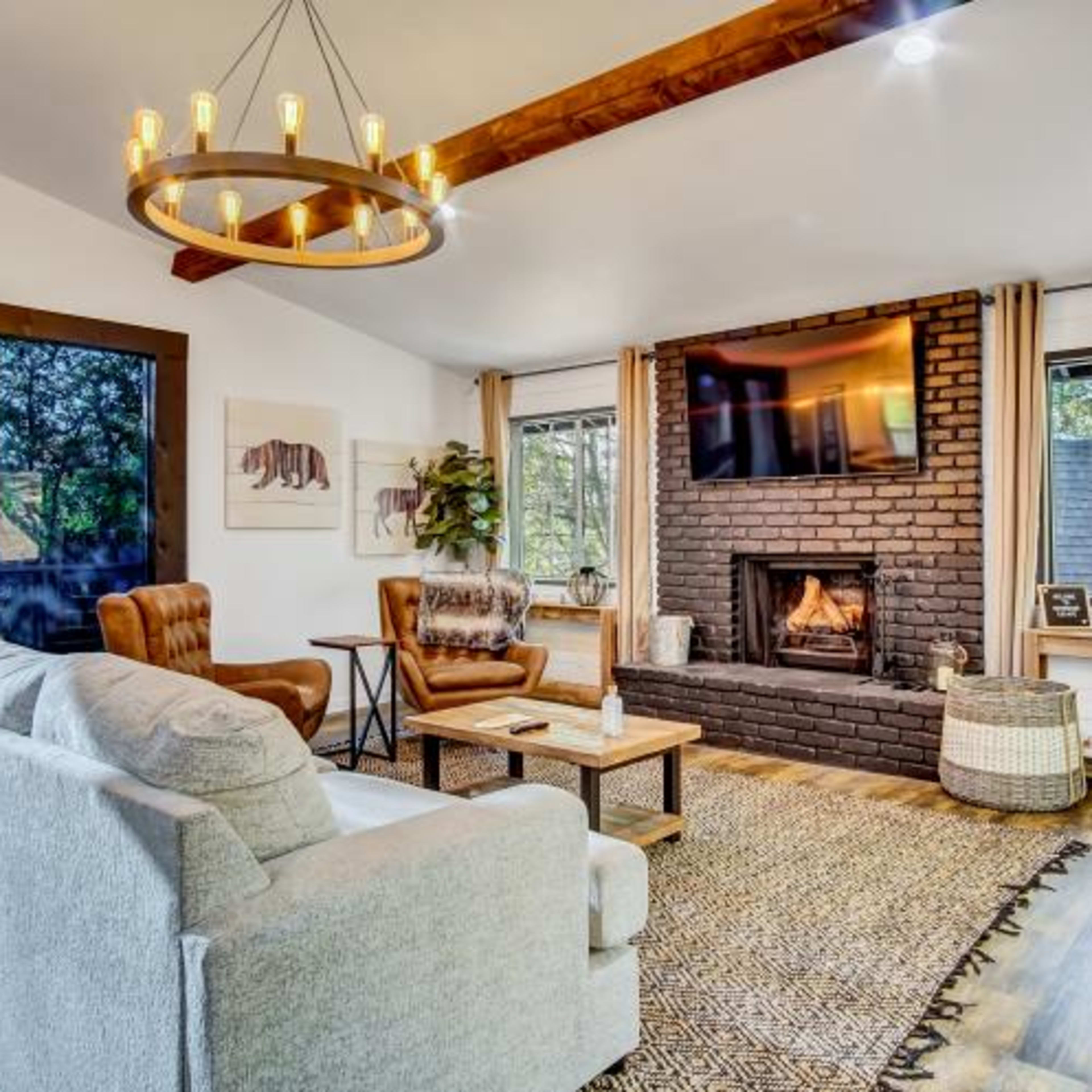A cozy living room with a fireplace, a television above it, a gray sofa, and a pair of armchairs, all accented by a patterned area rug and large windows.
