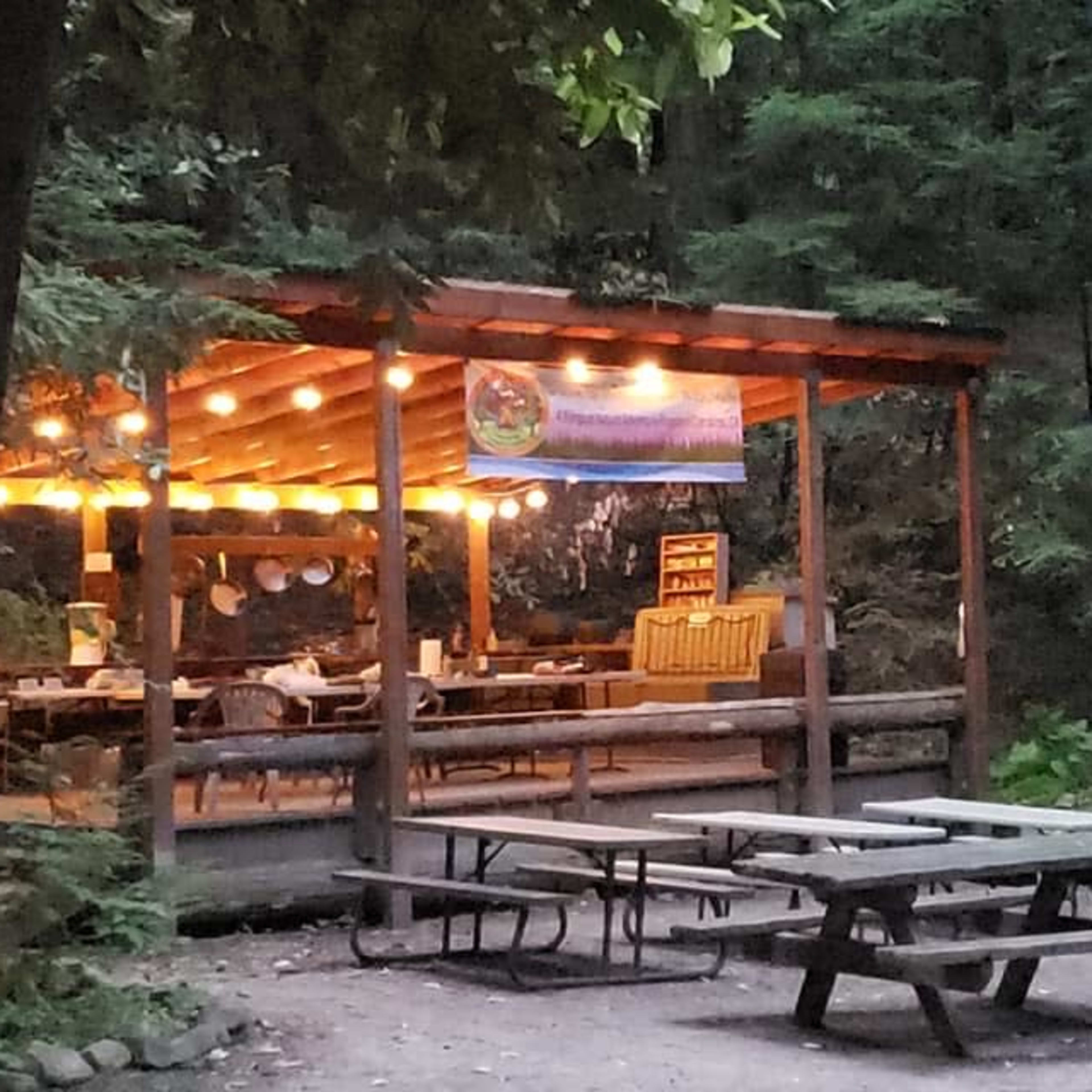 A wooden pavilion with string lights and picnic tables stands in a forested area, next to a staircase leading up to a wooded path.