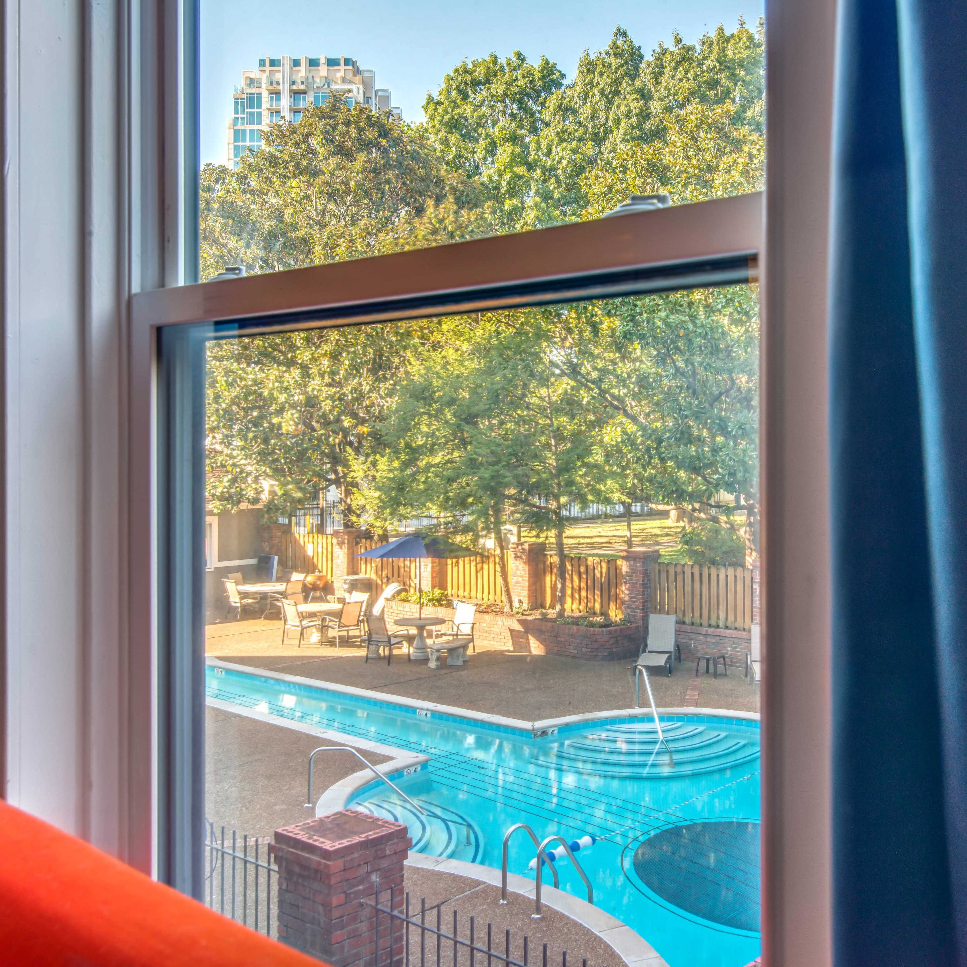 A window frame with a view of a swimming pool surrounded by trees and patio furniture.