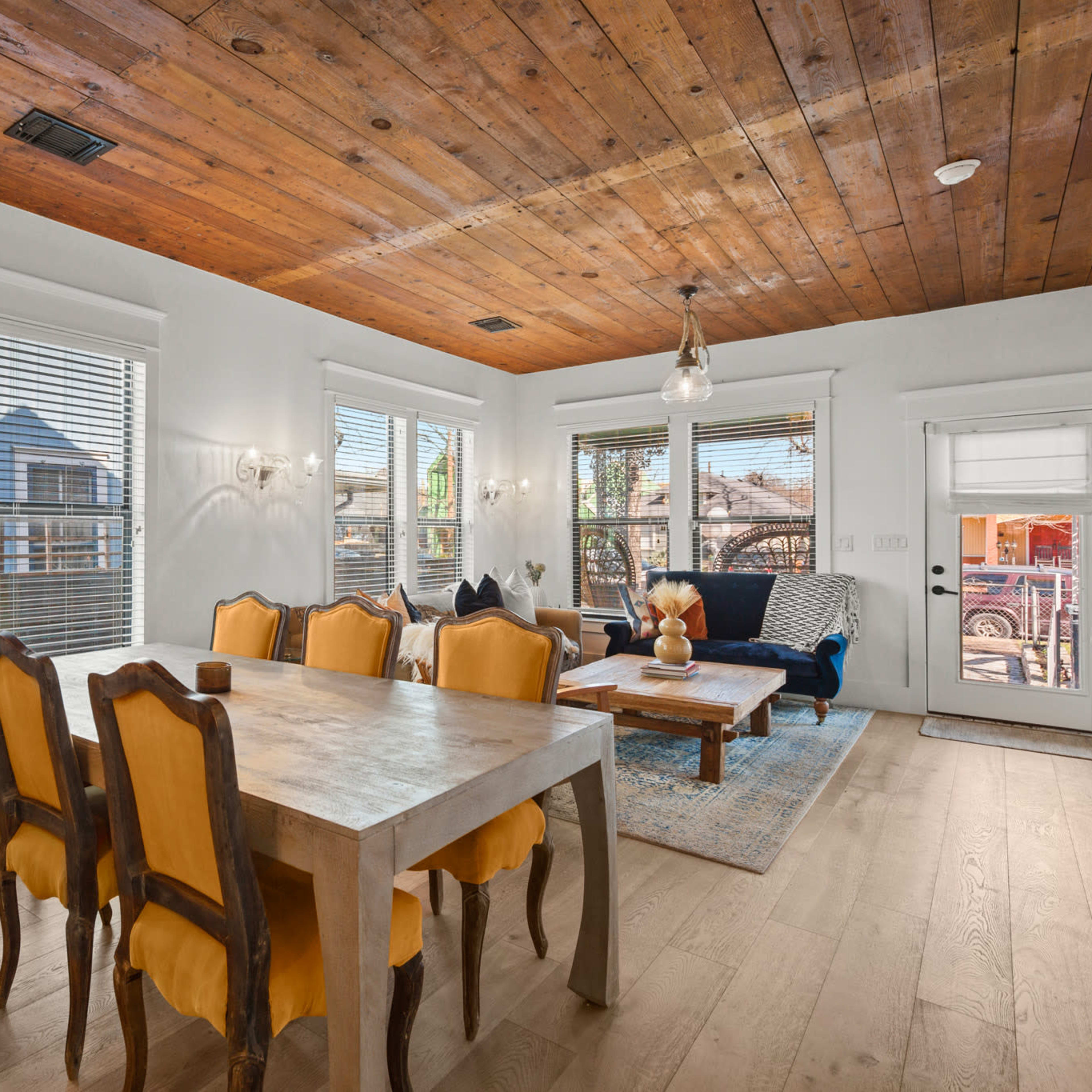 A dining area features a wooden table surrounded by yellow upholstered chairs, with large windows providing natural light and a view of the outdoors.