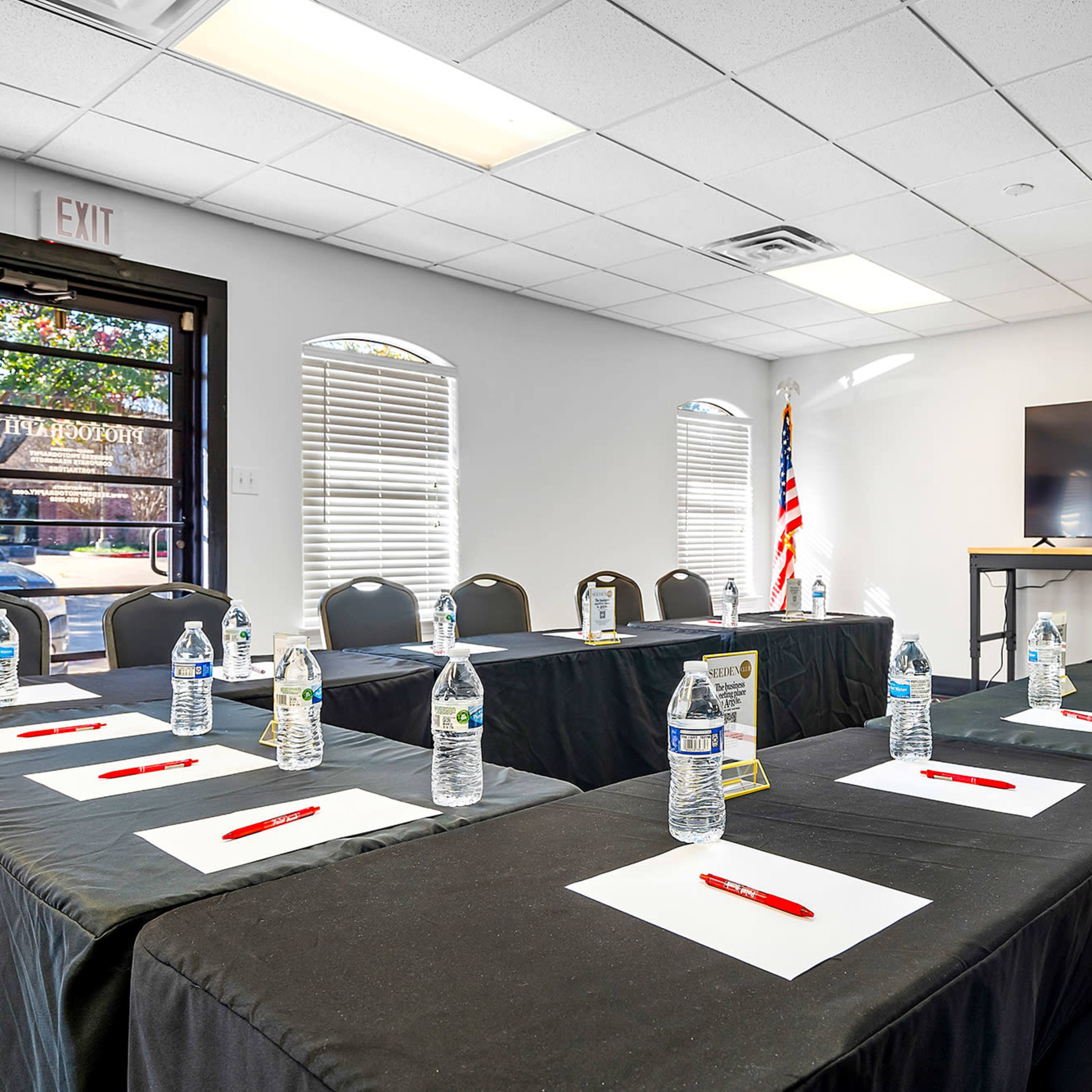 A conference room is set up with black tables, chairs, water bottles, and red pens, facing a large screen and an American flag.