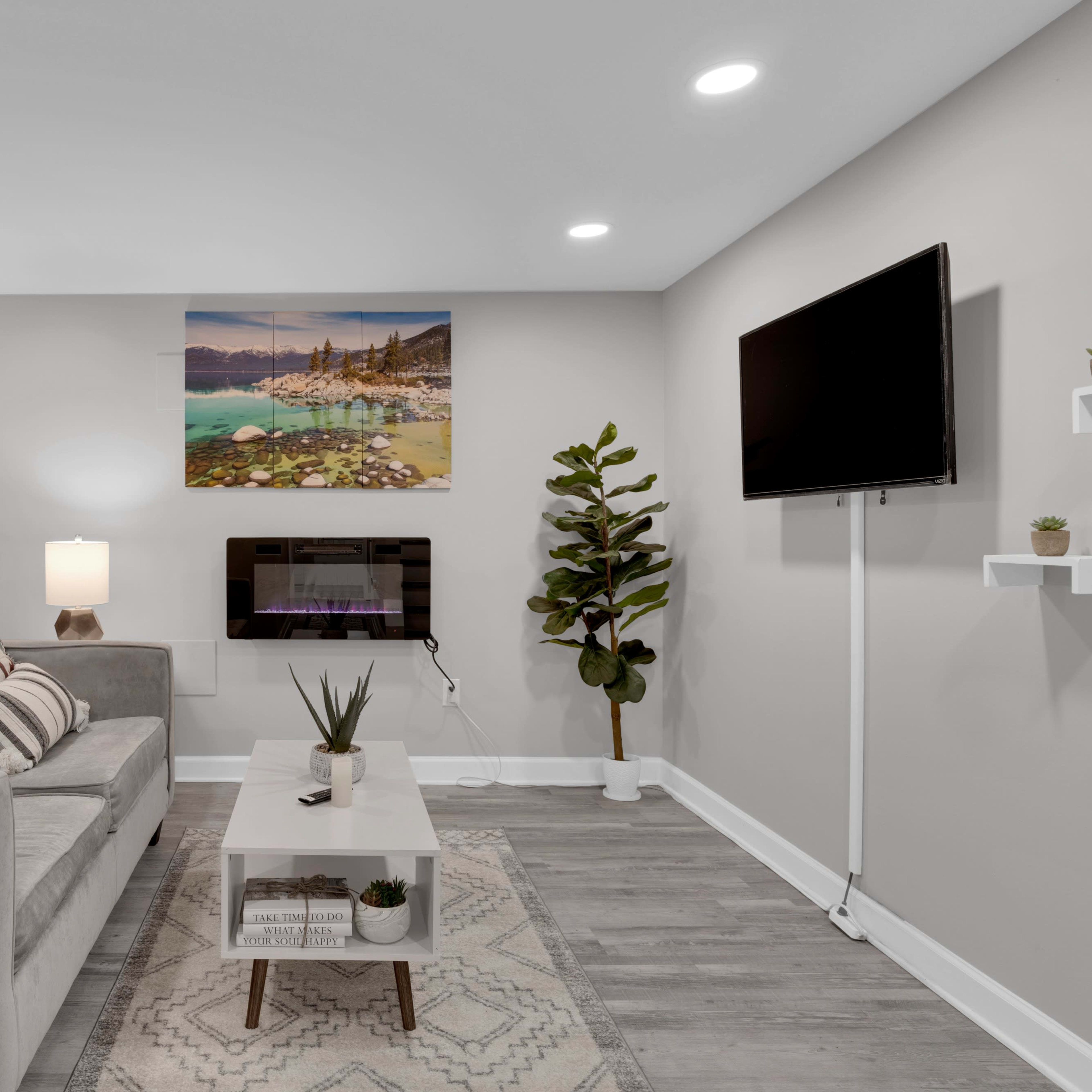 A modern living room with a light gray sofa, a wall-mounted TV, and a coffee table on a patterned rug, flanked by decorative plants.