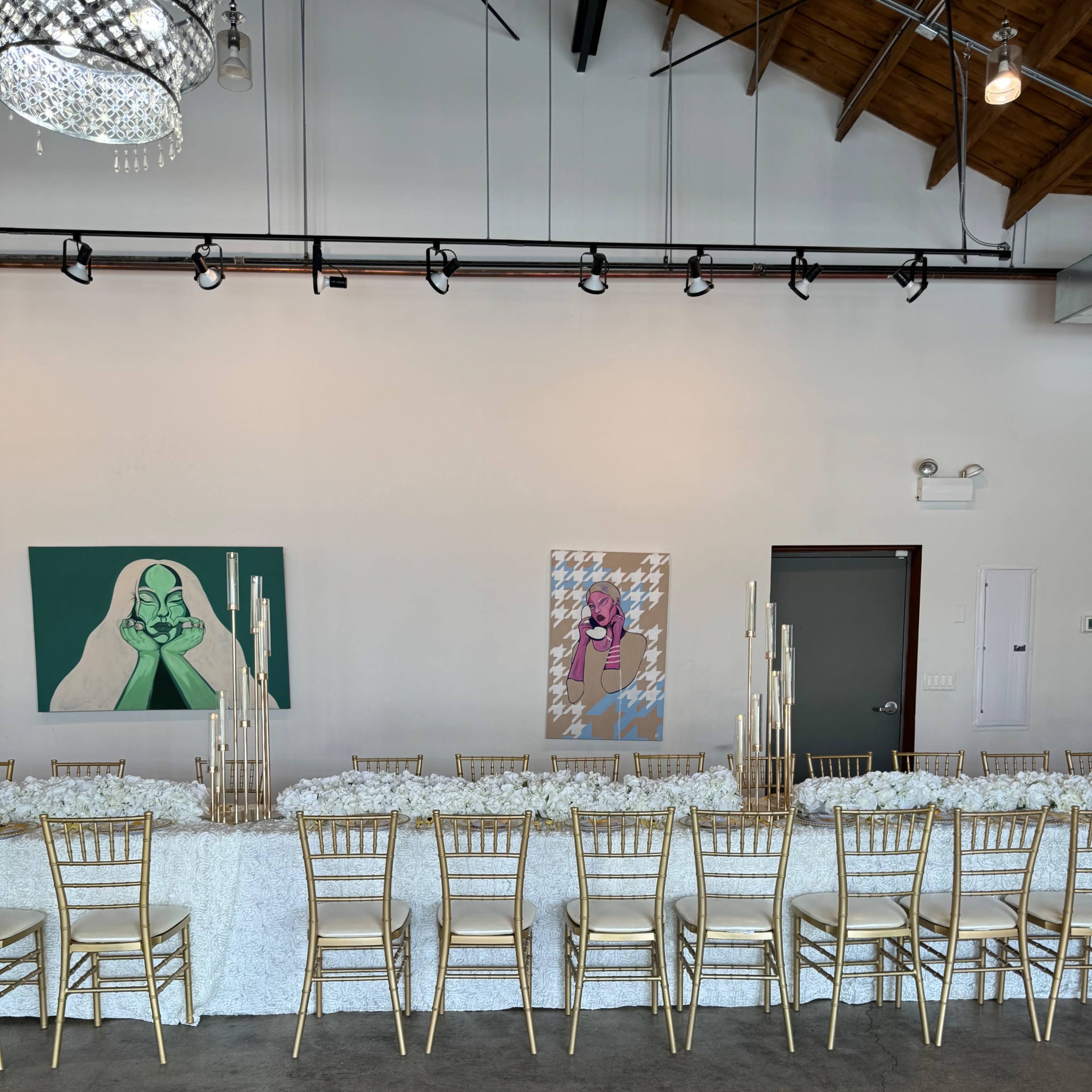 A long banquet table with gold chairs is set up in a modern interior featuring two colorful artworks on the walls.