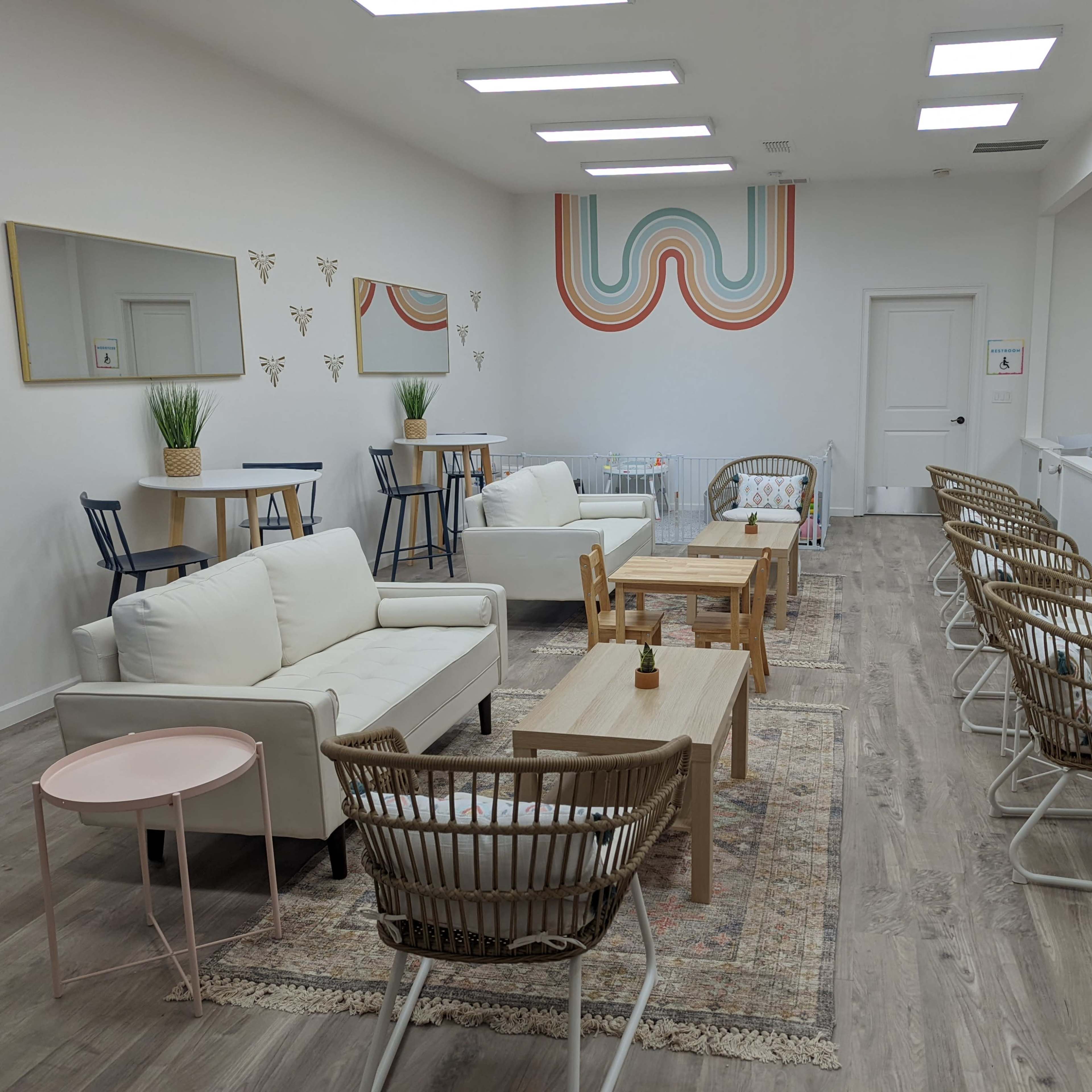 The scene features a spacious, well-lit lounge area with white couches, wooden tables, and rattan chairs arranged on a wooden floor, complemented by plants and decorative wall art.