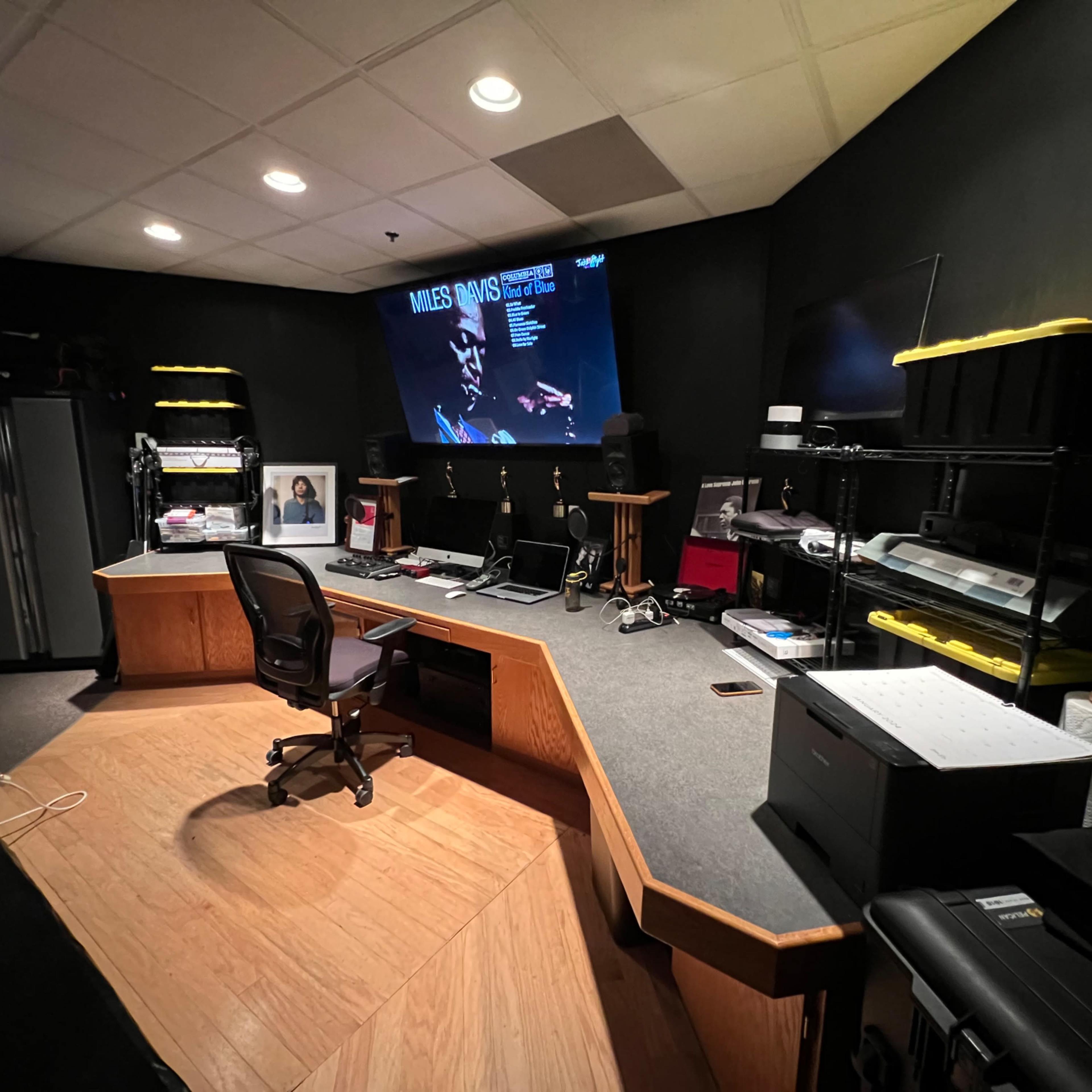 A spacious audio-visual production studio features a large screen displaying information, surrounded by various equipment, a desk with a chair, and storage shelves.
