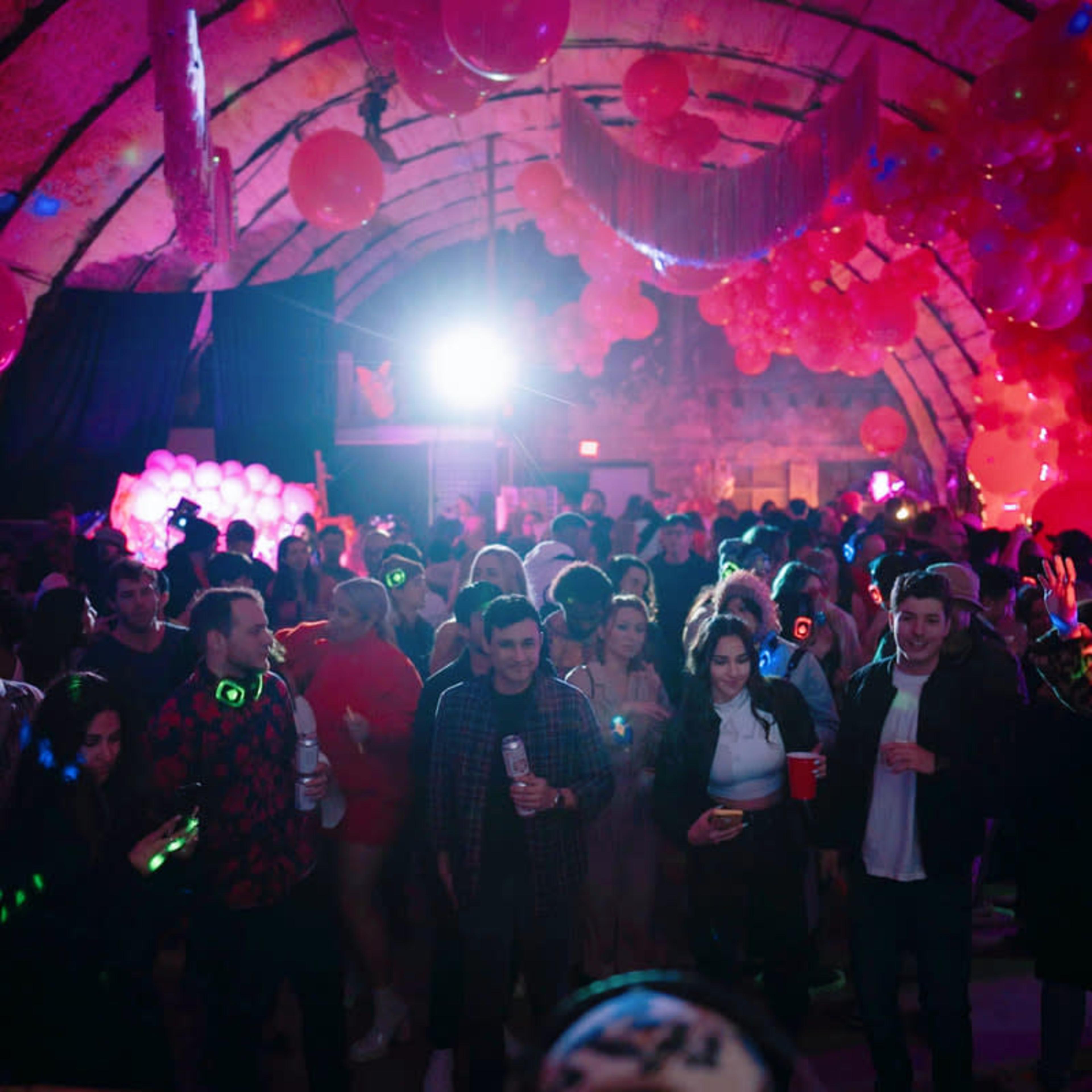 A crowded party scene is illuminated by colorful lights and filled with people holding drinks and wearing glow sticks.