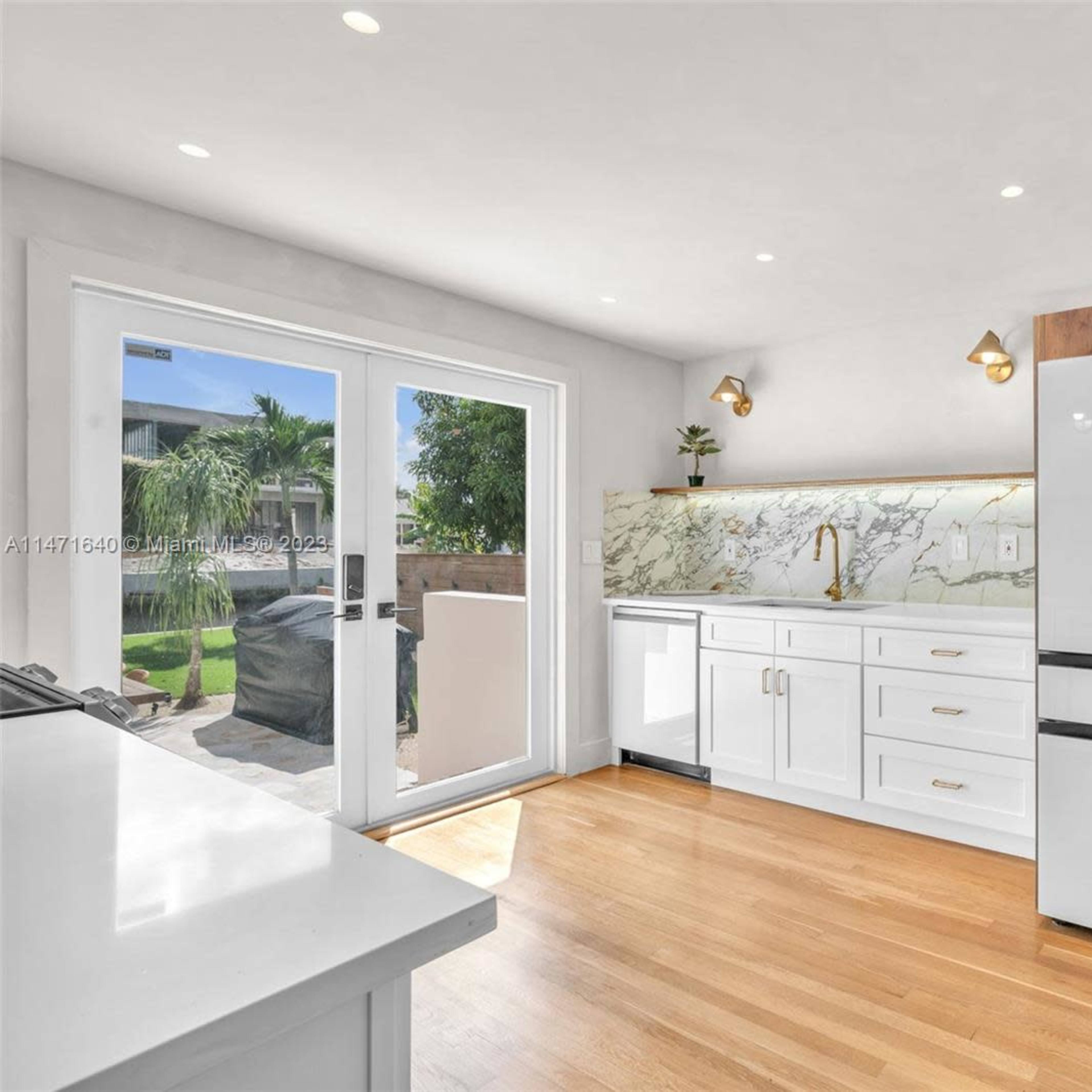 A modern kitchen with white cabinetry, a large fridge with wooden accents, and a marble backsplash, illuminated by natural light from a nearby door.