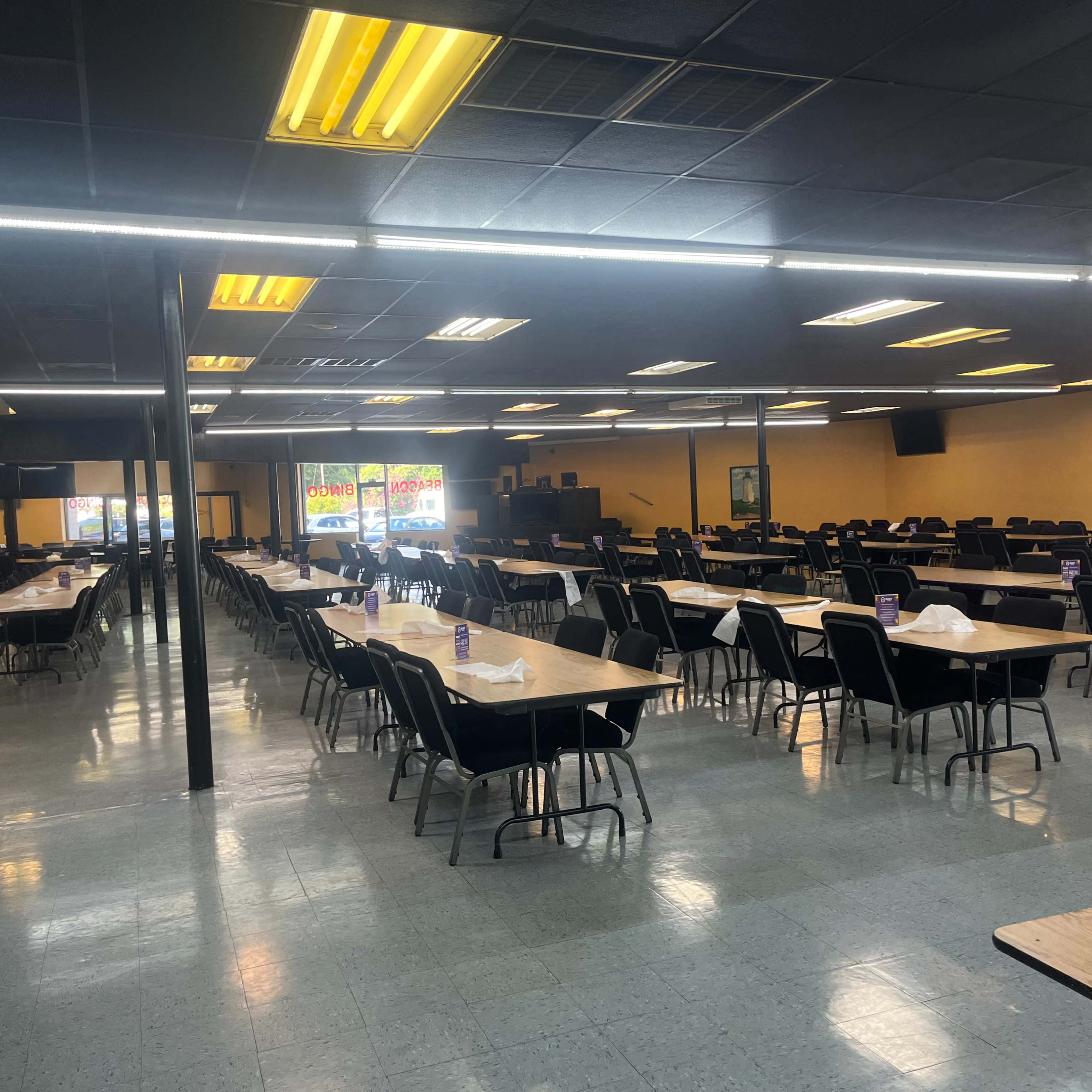 A large, empty dining area with multiple rows of tables and chairs, illuminated by overhead lights.