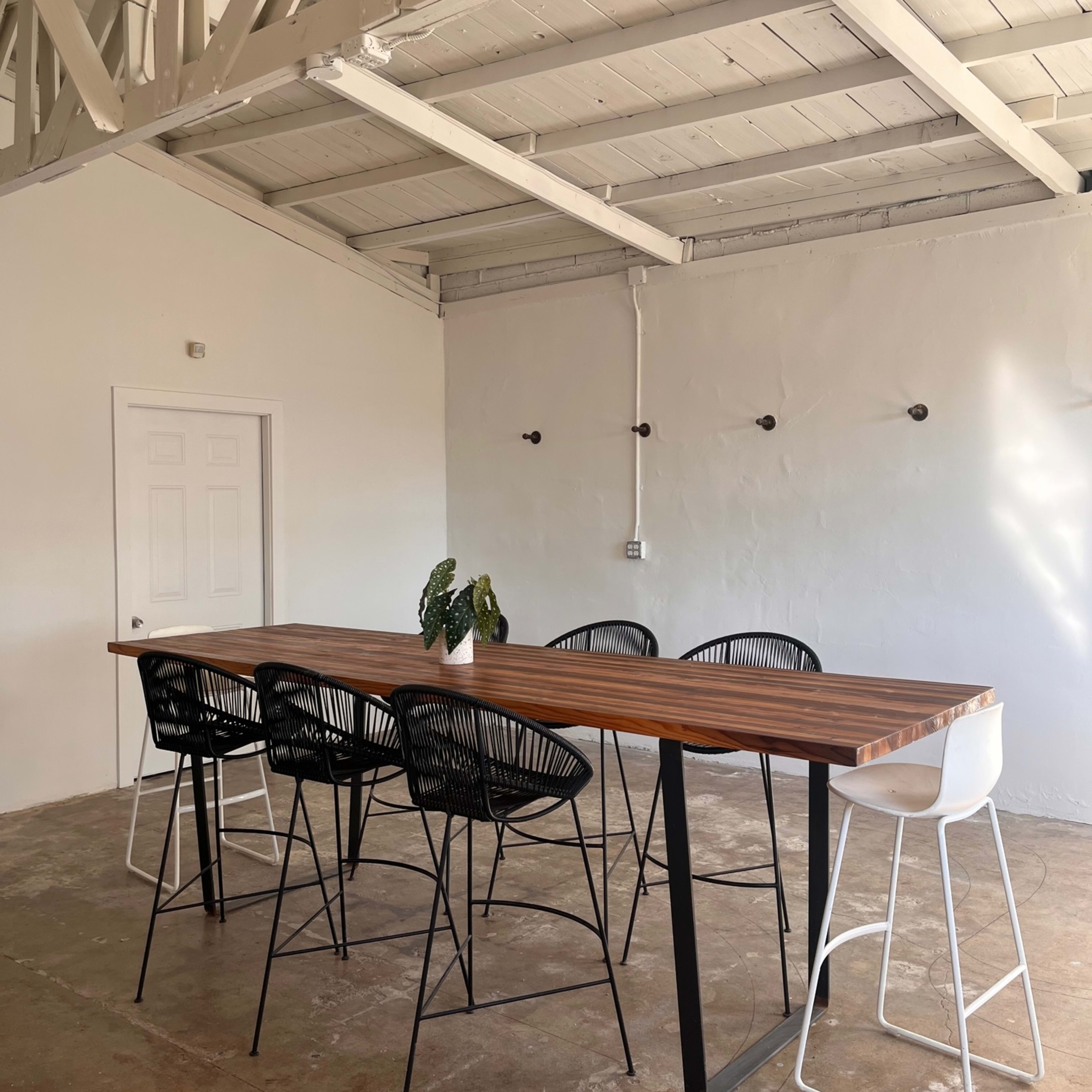A long wooden table with black and white high chairs is set in a minimalistic room with a white ceiling and walls.