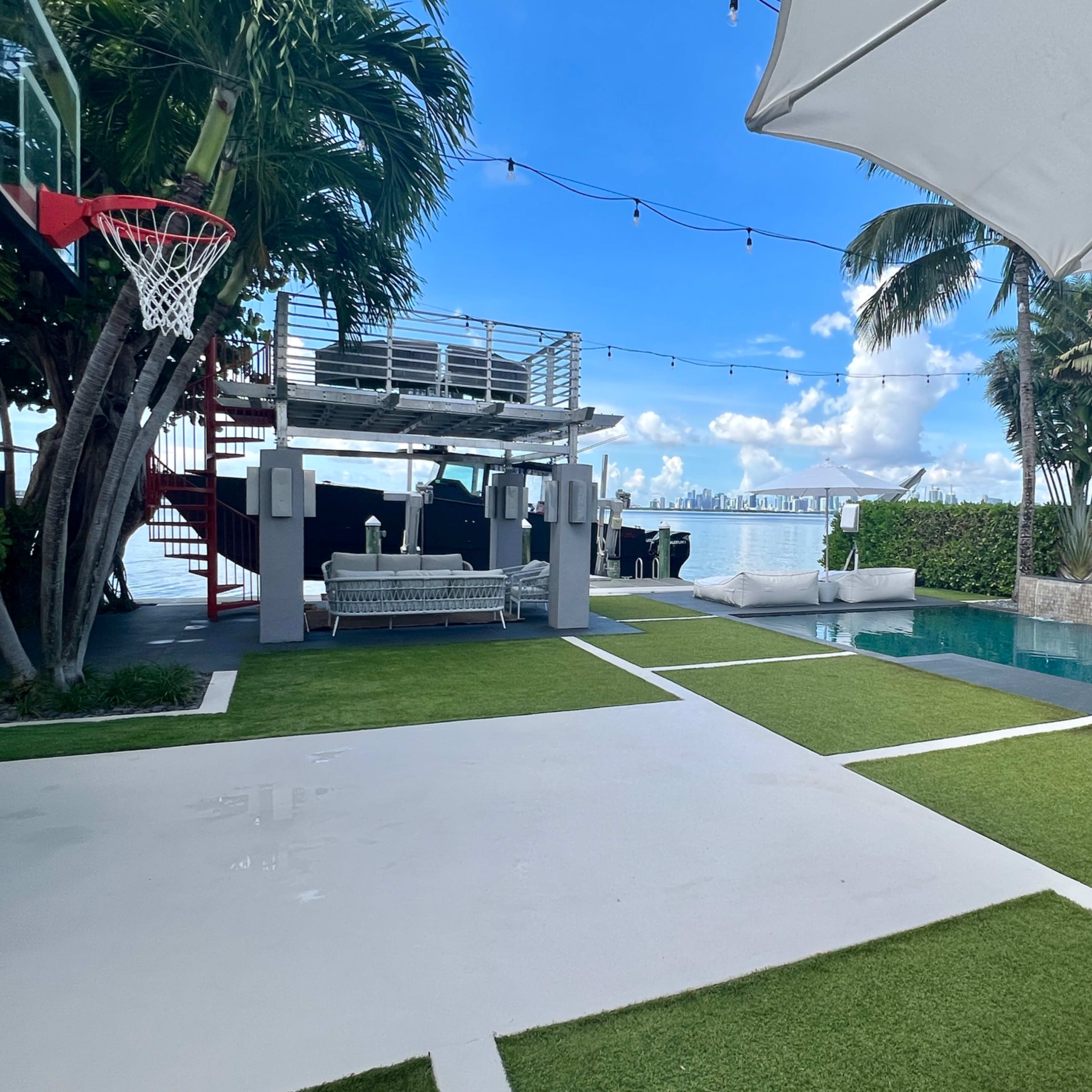 A landscaped outdoor area featuring a swimming pool, tropical plants, a basketball hoop, and a shaded seating area.