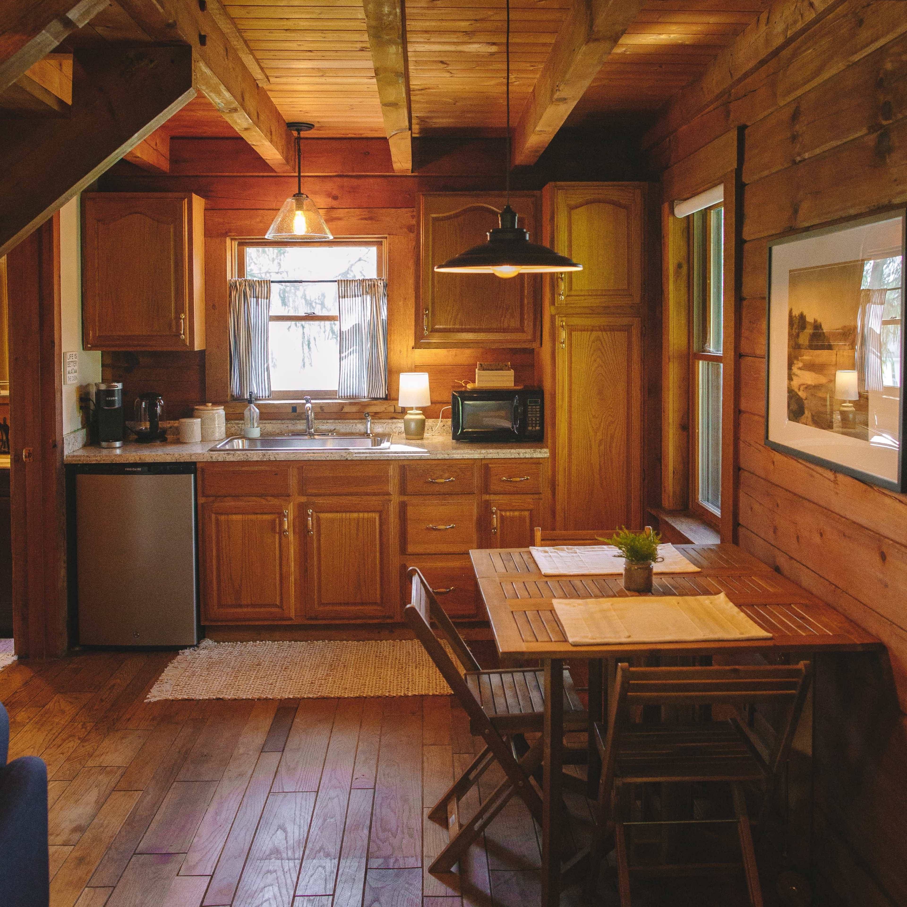 A wooden kitchen with cabinets, a dining table, and various appliances under warm lighting.