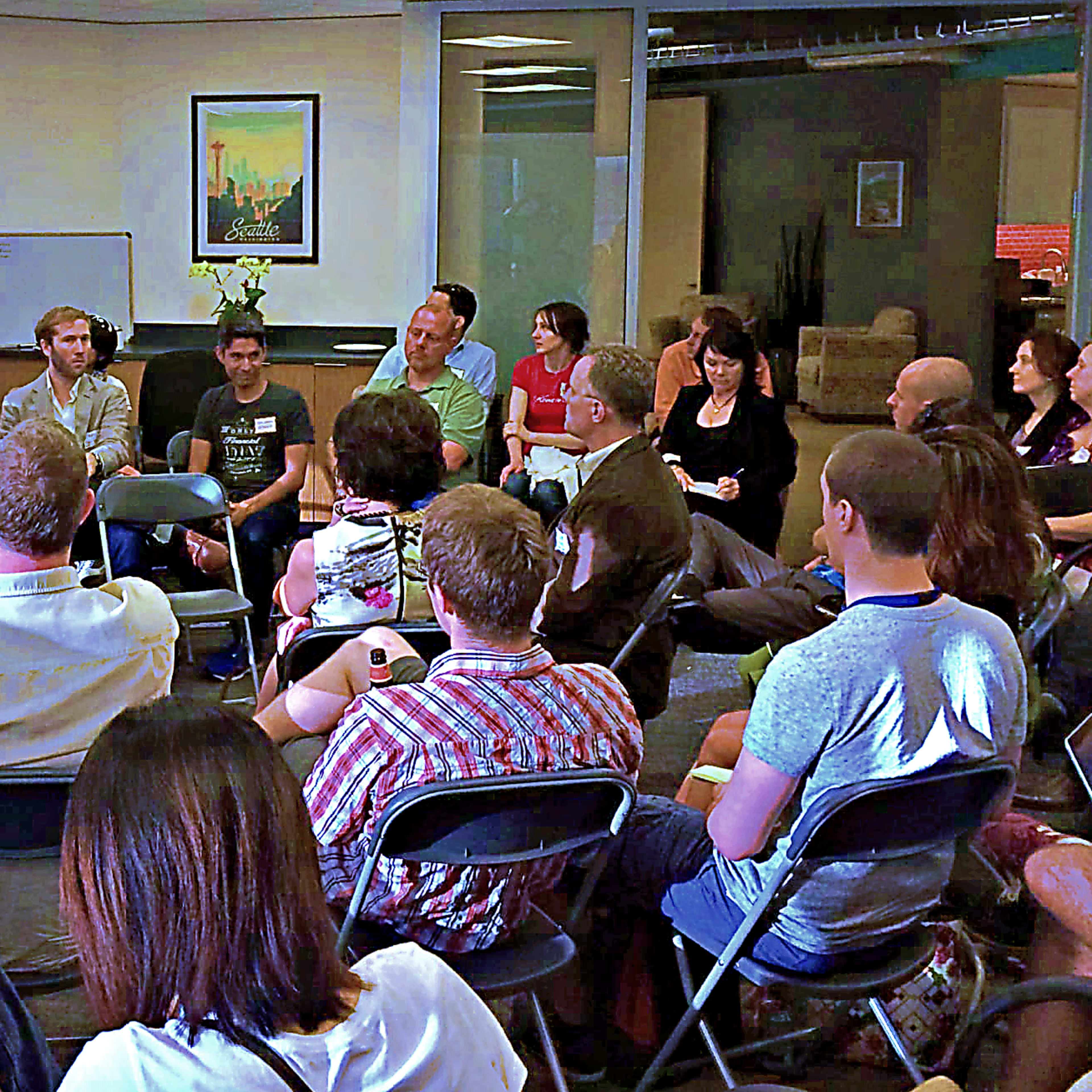 A diverse group of people sits in a semi-circle facing a speaker in a conference room setting.