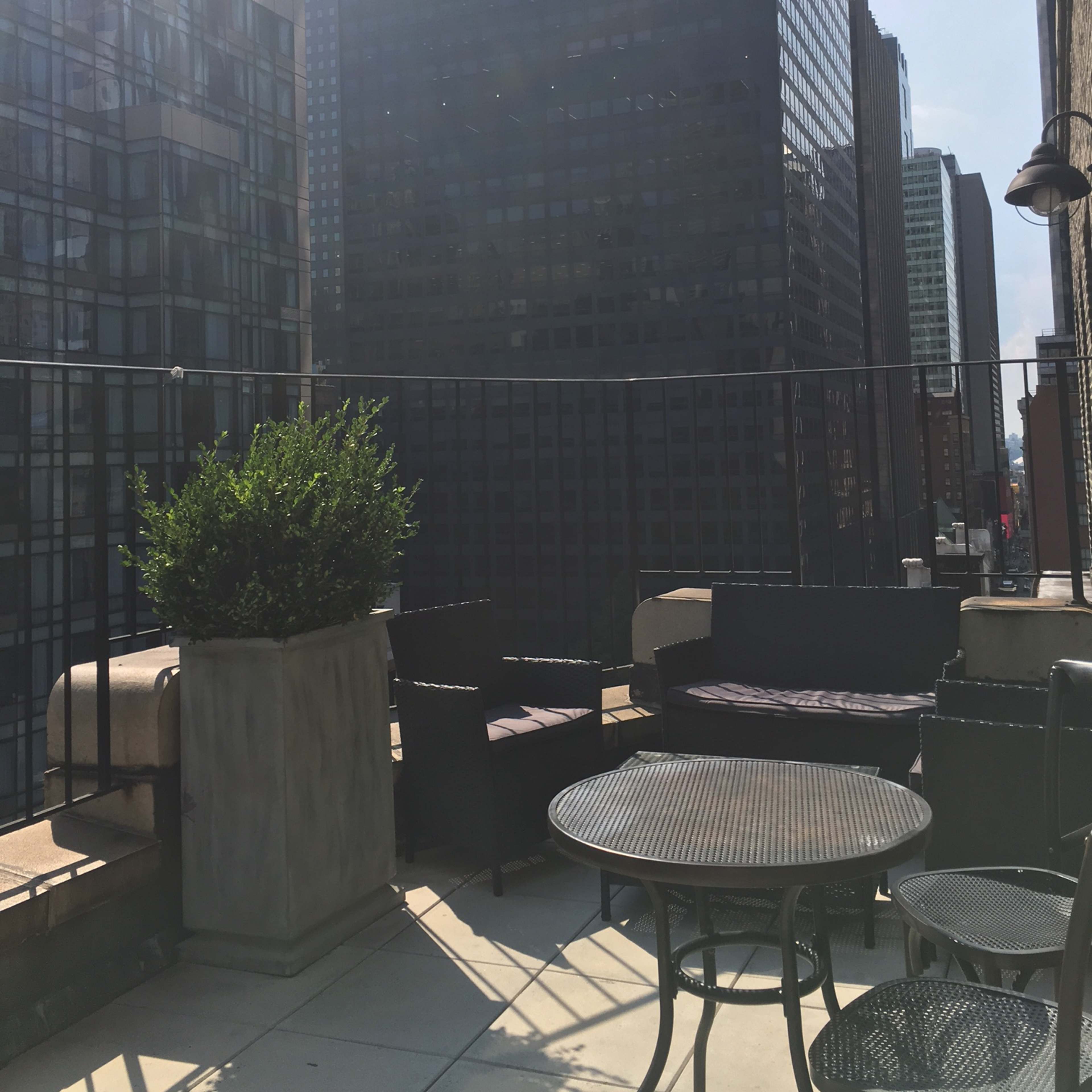 A rooftop terrace with modern furniture overlooking a cityscape of tall buildings under a partly cloudy sky.