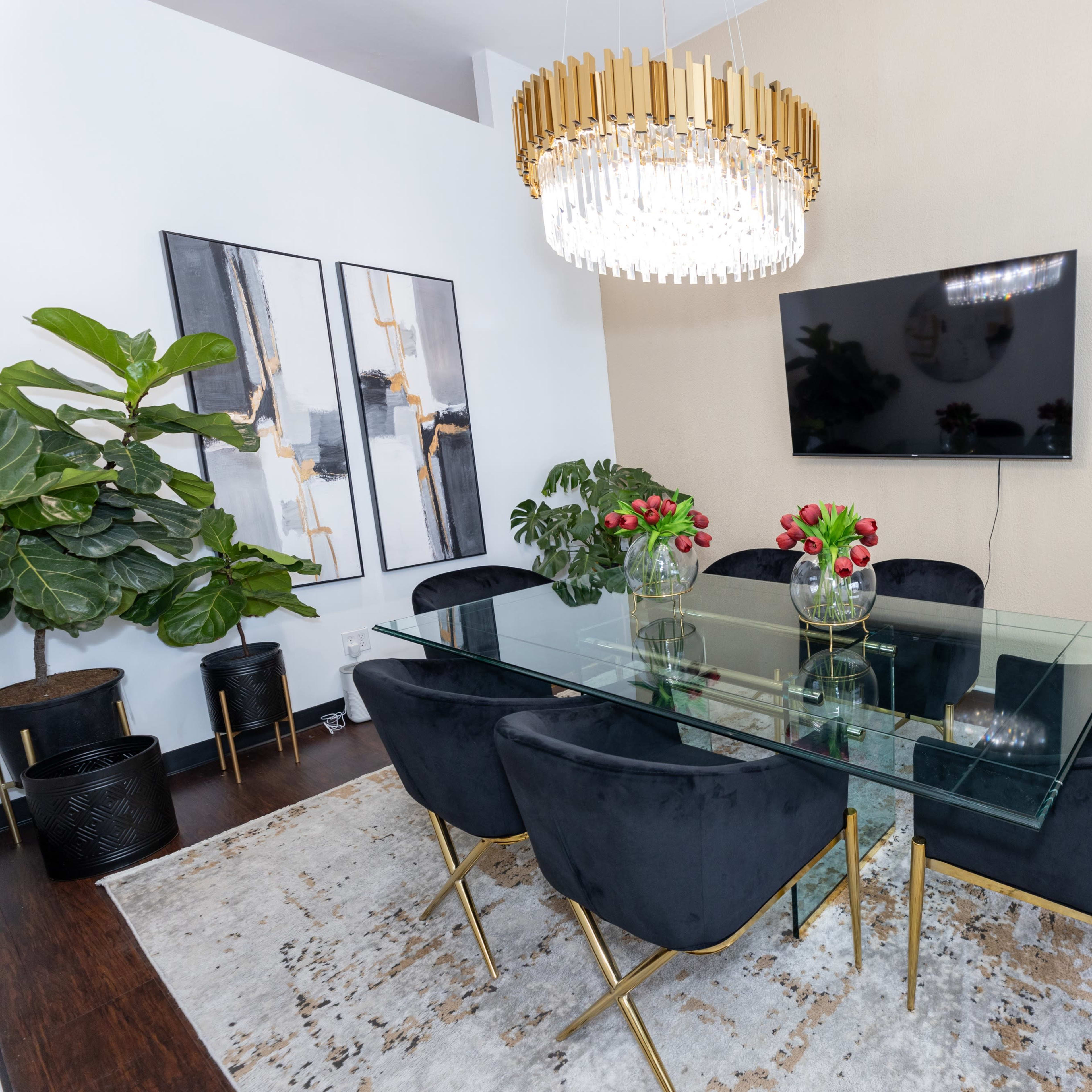 A modern dining room featuring a glass table surrounded by black chairs, a large chandelier overhead, and decorative plants along with wall art.