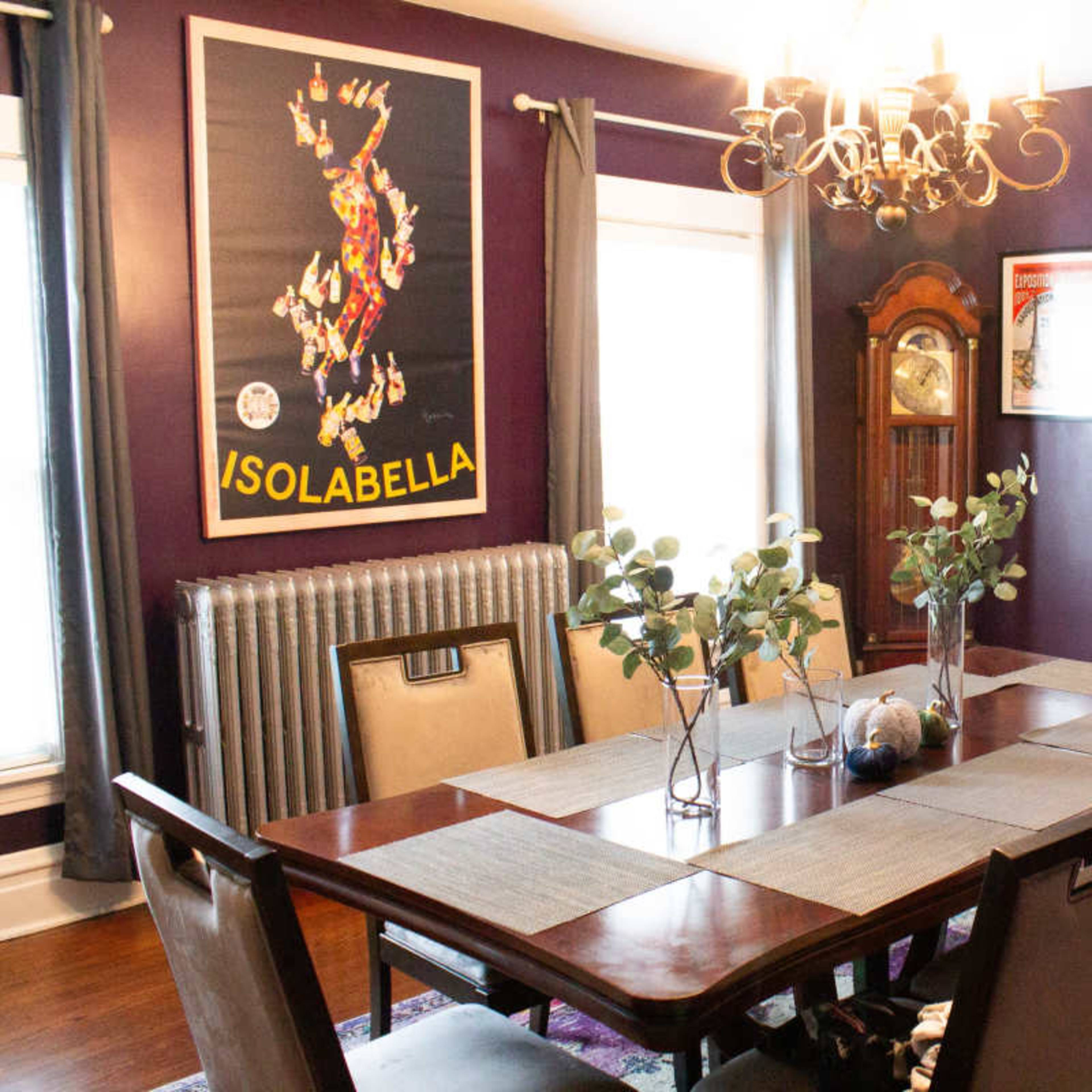 A dining room features a long wooden table with chairs surrounded by purple walls, a chandelier overhead, and a vintage poster on display.