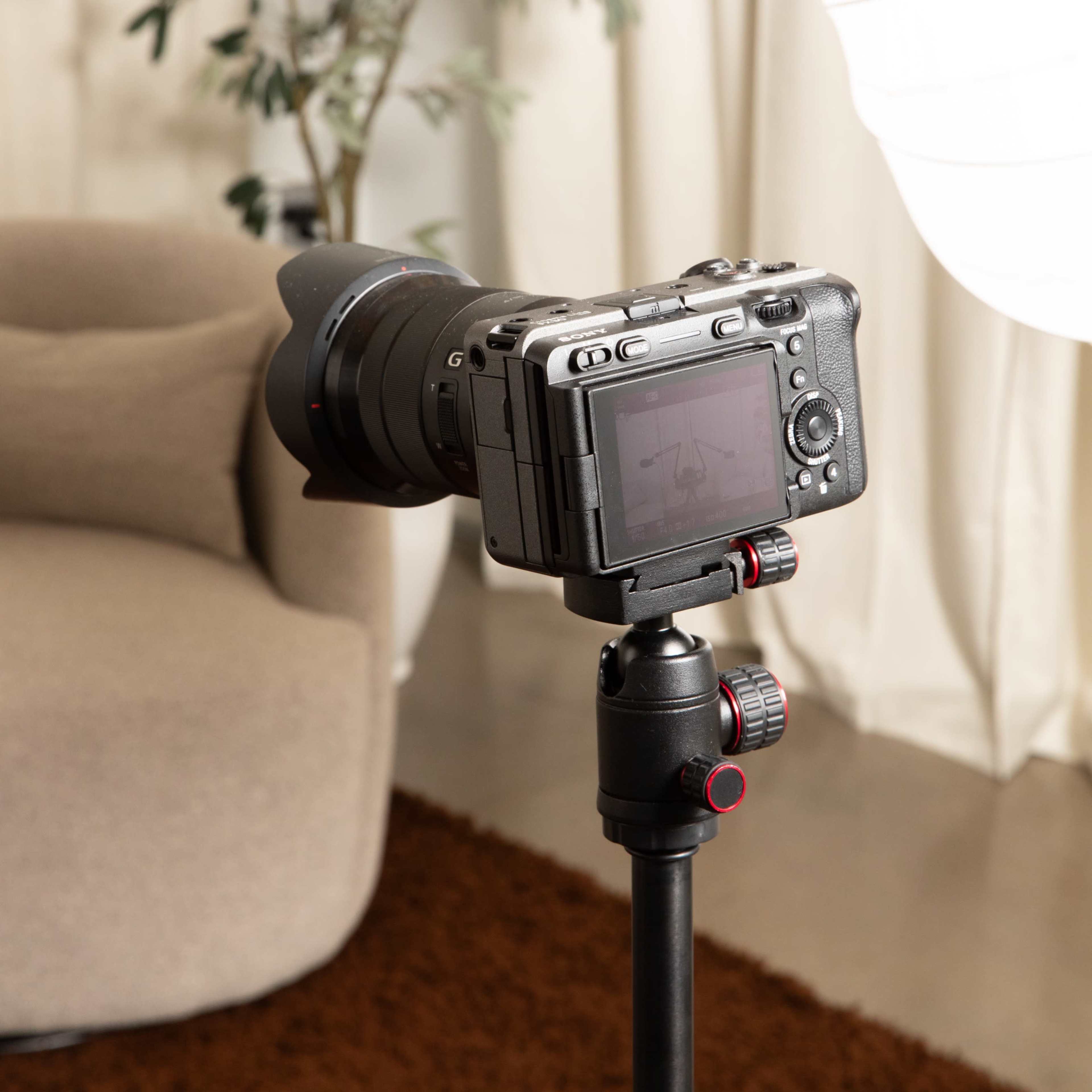 A camera is positioned on a tripod facing a soft light source with a large white lamp, near a beige sofa and a potted plant.