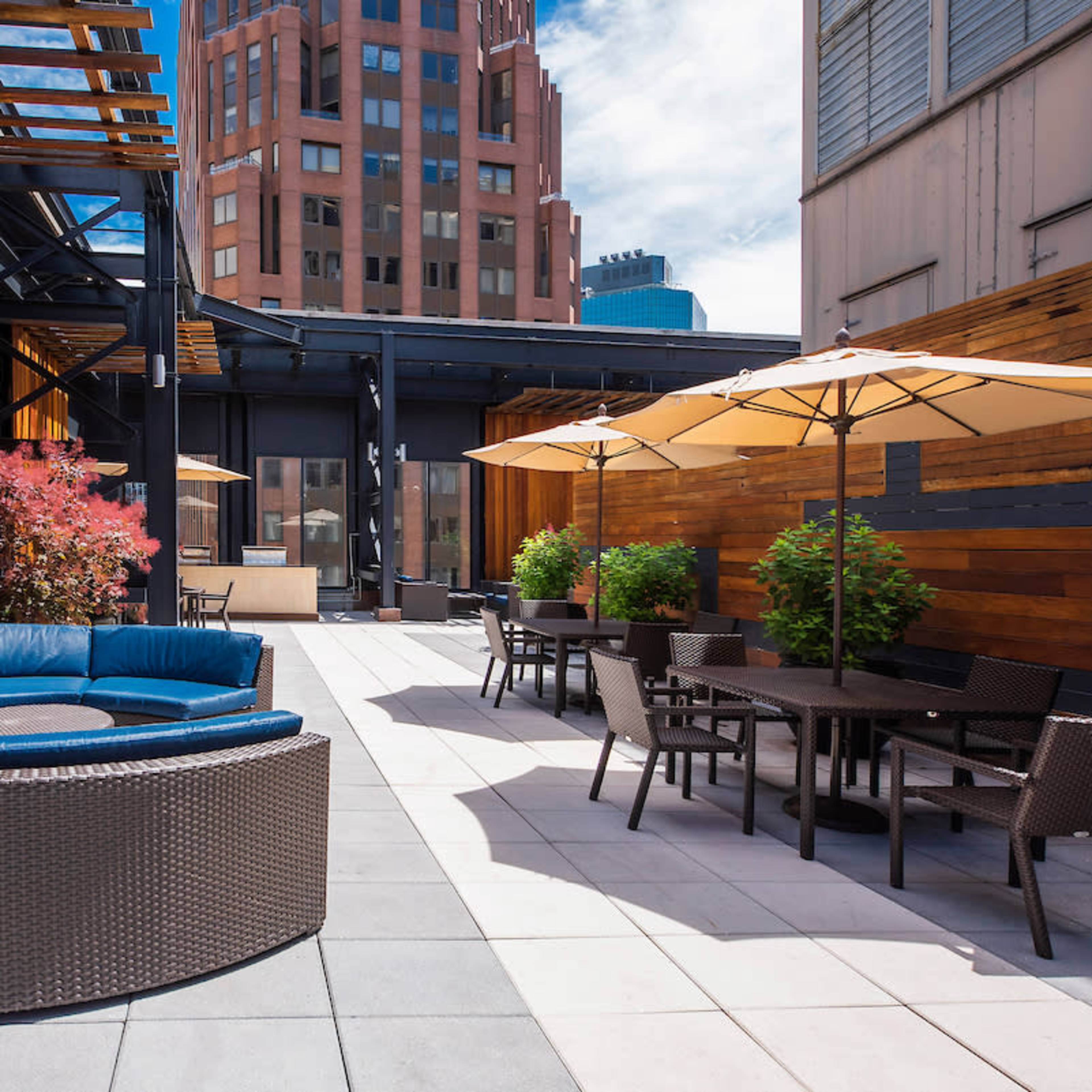 A rooftop patio featuring a blue circular seating arrangement, umbrella-covered tables, and wooden paneling with pink foliage.