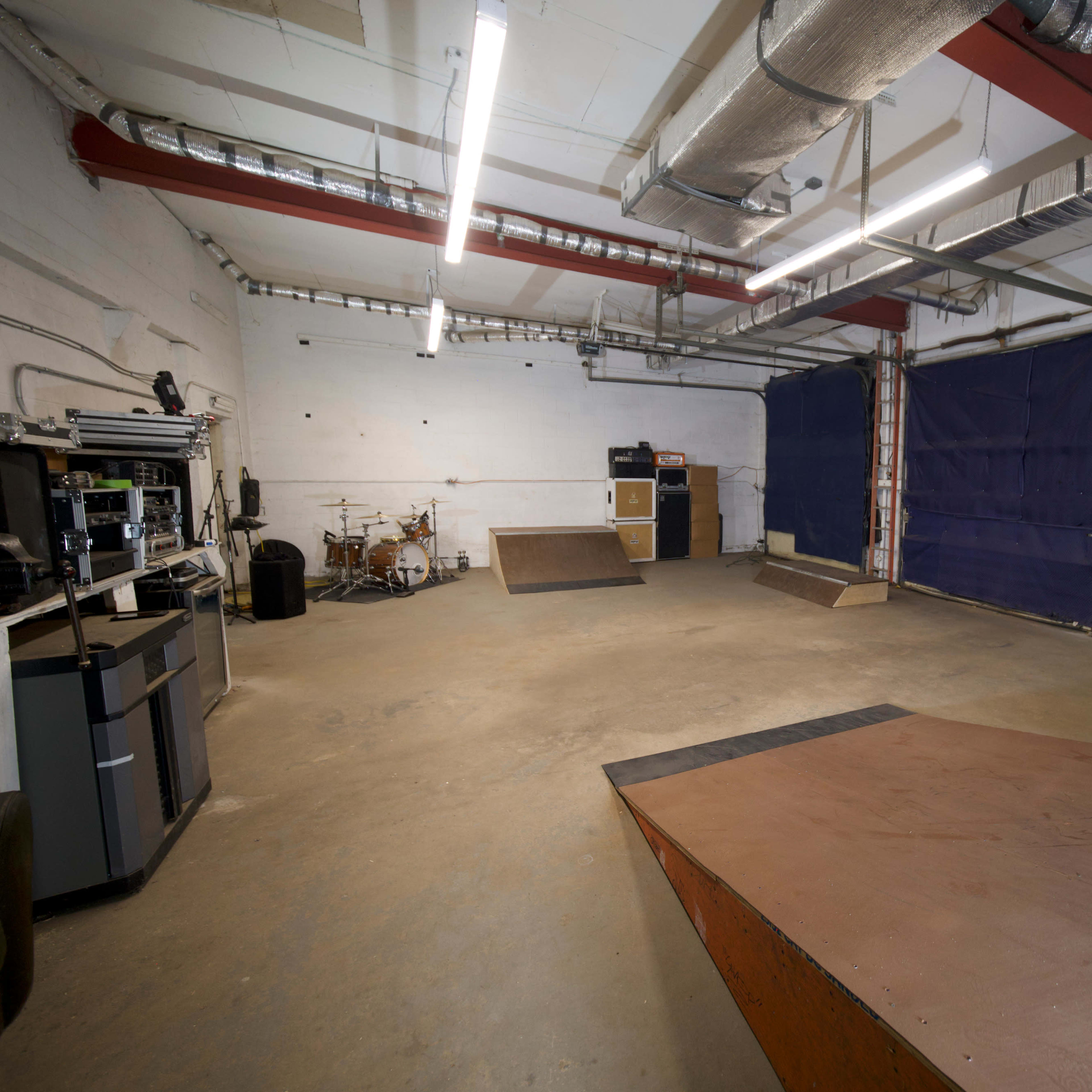 An empty indoor space with two ramps, a drum set, and exposed beams, illuminated by overhead fluorescent lights.