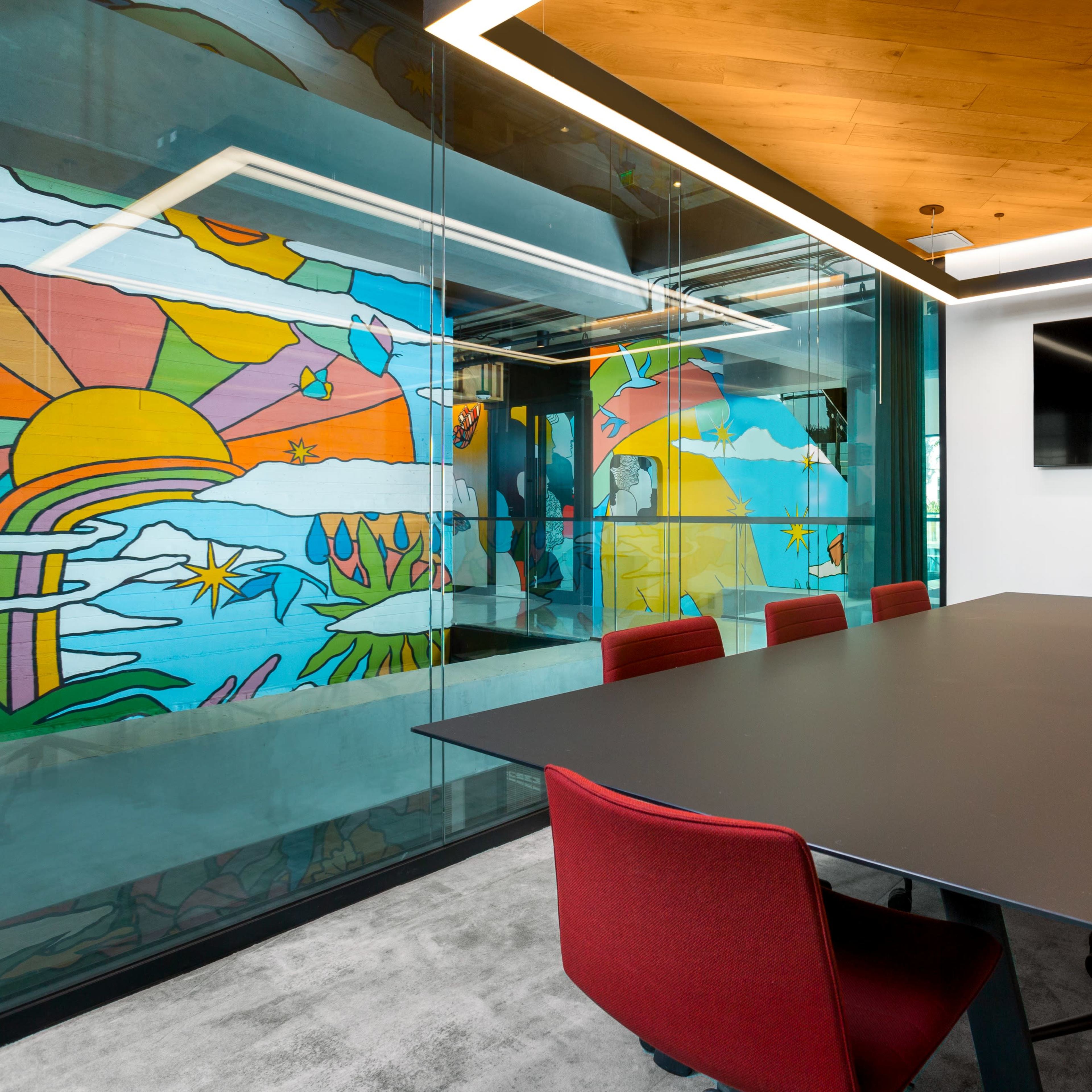 A modern conference room features a large black table surrounded by red chairs, with glass walls displaying a colorful mural and a mounted television.