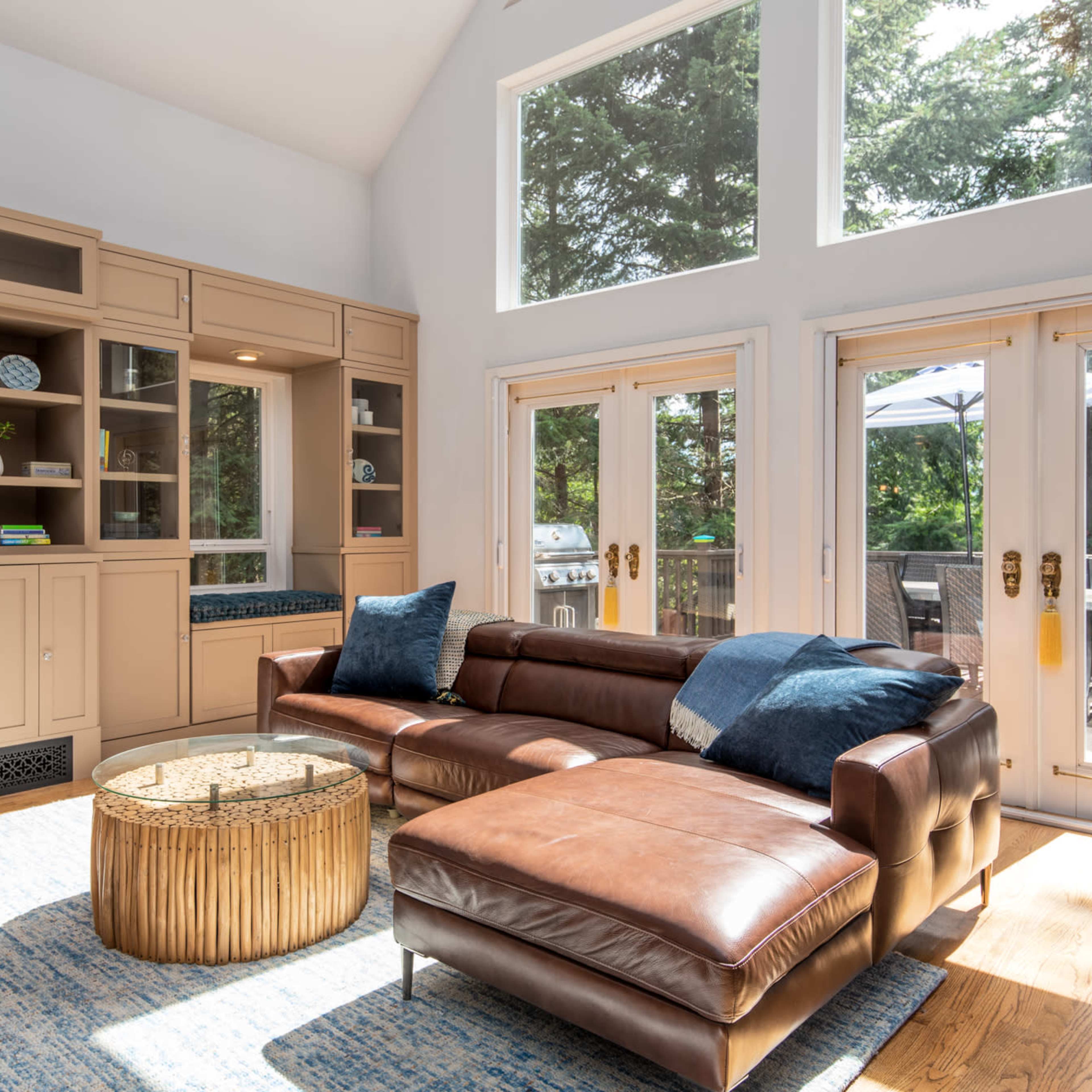 A sunlit living room features a brown leather sectional sofa, a circular gold coffee table, built-in cabinetry, and large windows overlooking a deck.