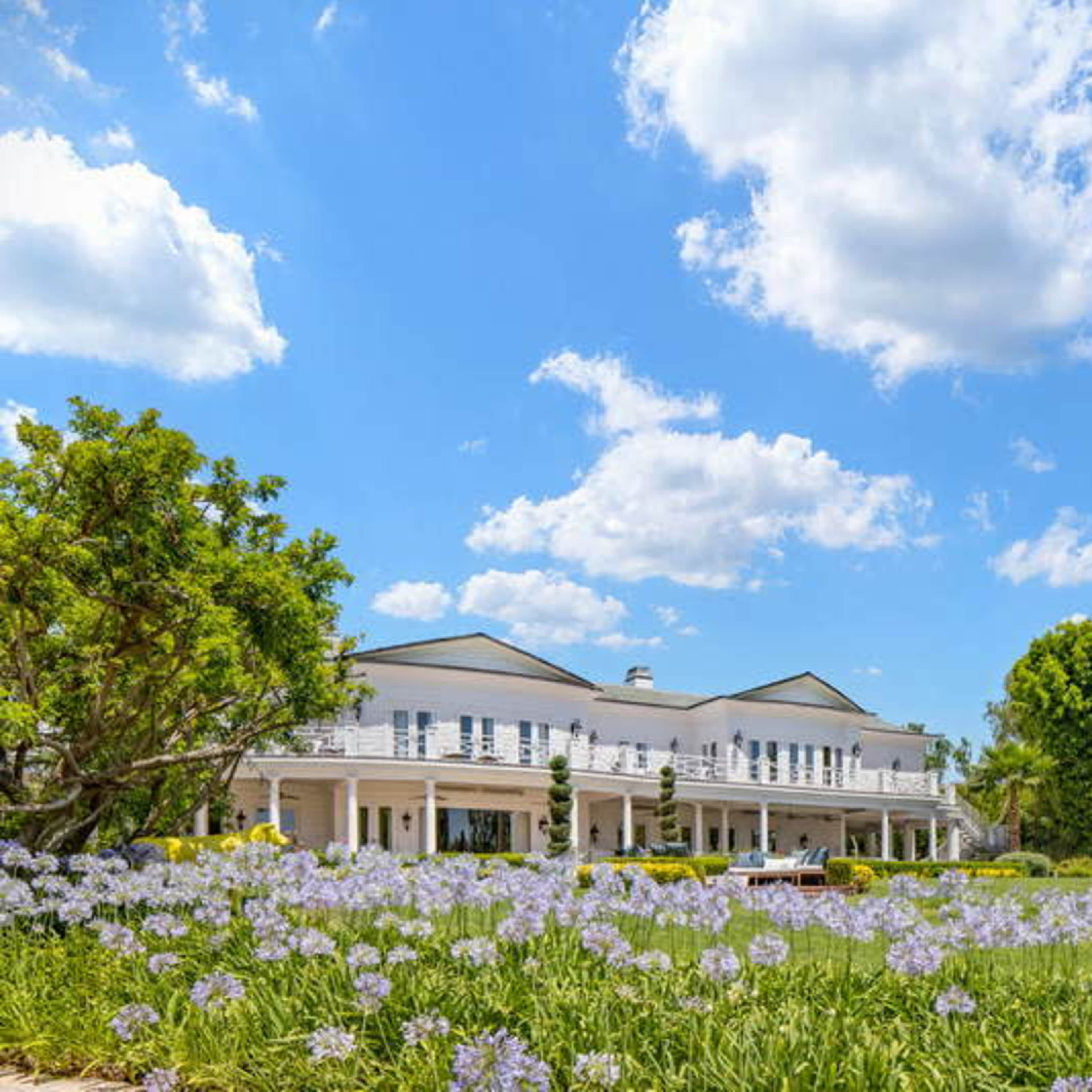 A large white mansion with a wraparound porch is set against a backdrop of blue sky and scattered clouds, surrounded by green lawns and blooming flowers.