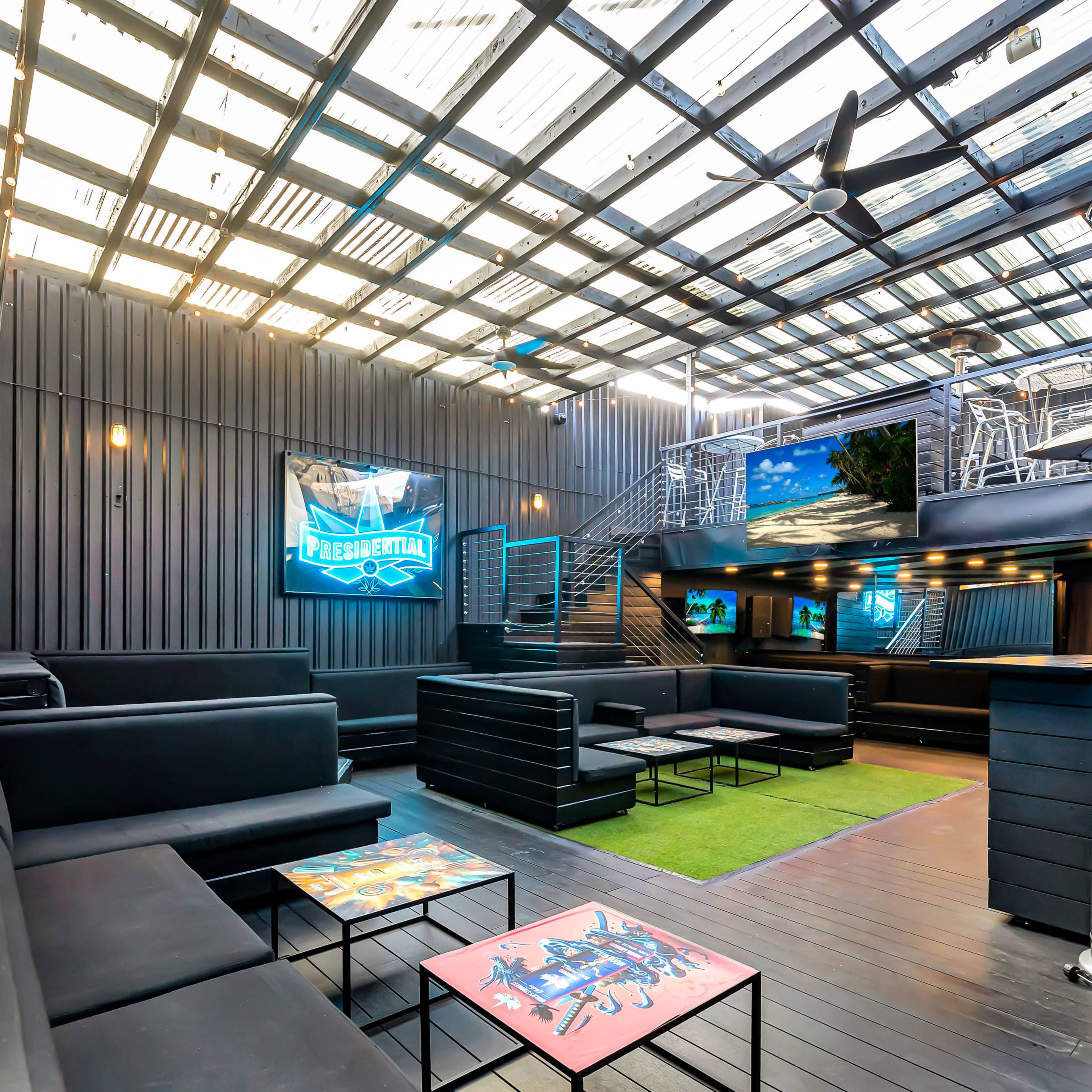 A spacious rooftop lounge area with black seating and flat-screen TVs on the walls, illuminated by overhead lighting and a glass ceiling.