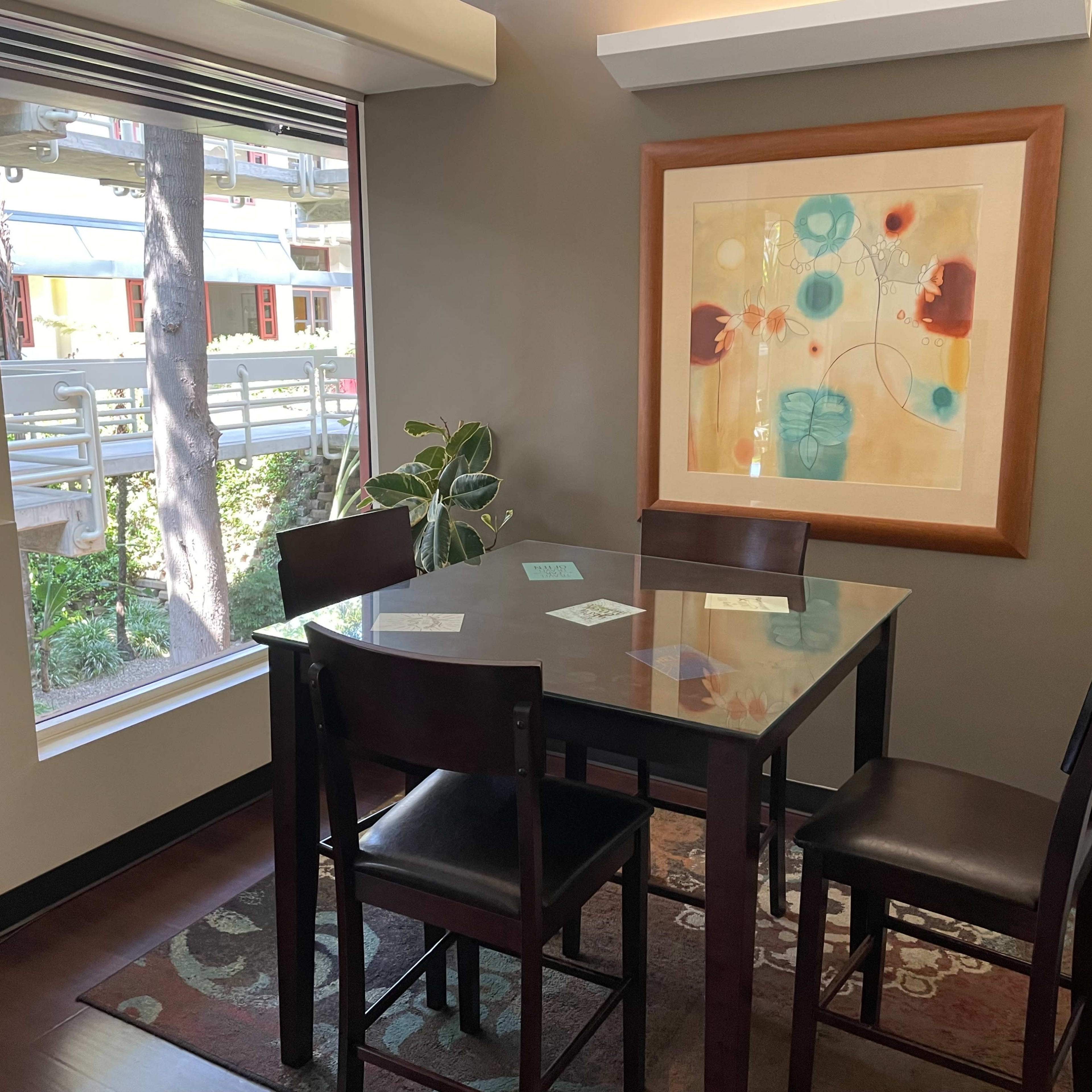 A dining area features a glass table surrounded by four chairs, with a framed artwork on the wall and a view of greenery outside through a large window.