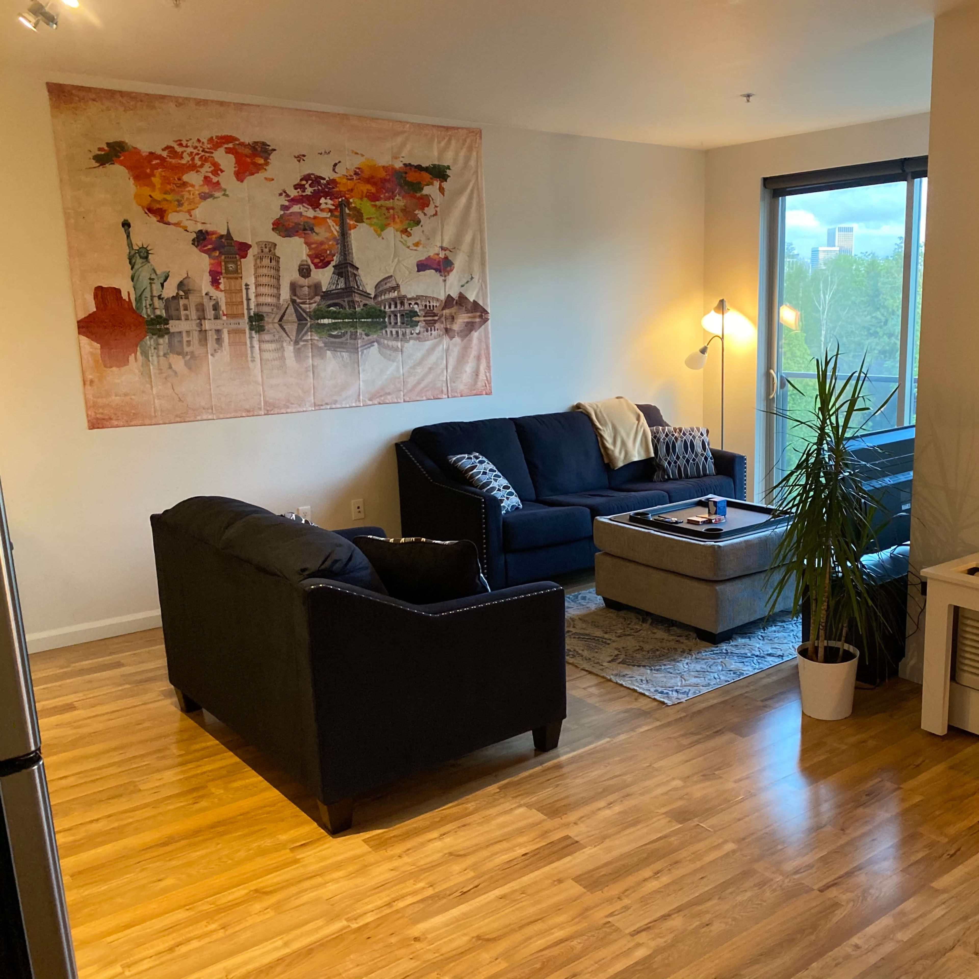 A modern living room featuring a map-themed tapestry on the wall, two black sofas, a coffee table, and large windows with natural light.
