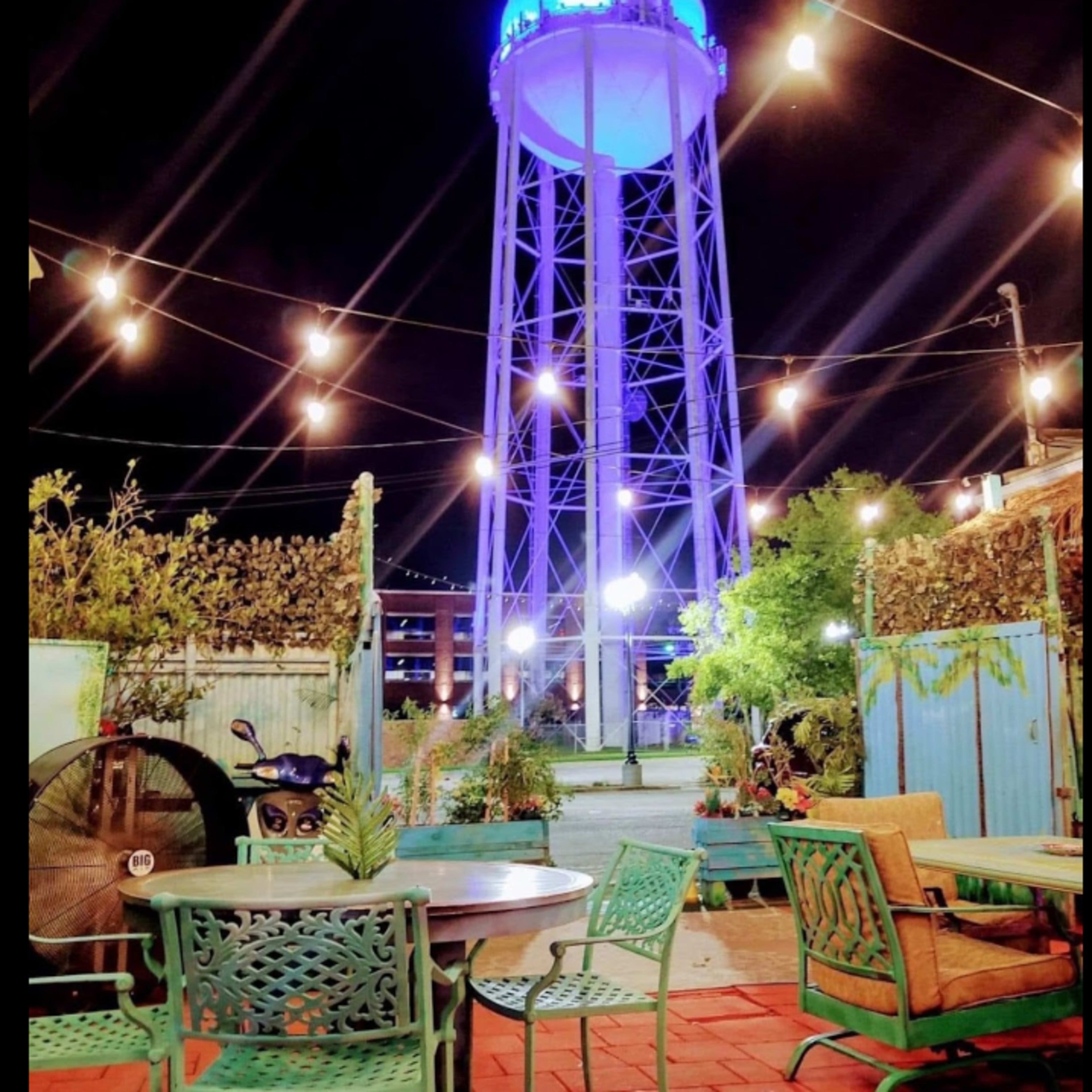 A brightly lit water tower stands illuminated in blue amidst outdoor seating and string lights in an urban courtyard at night.