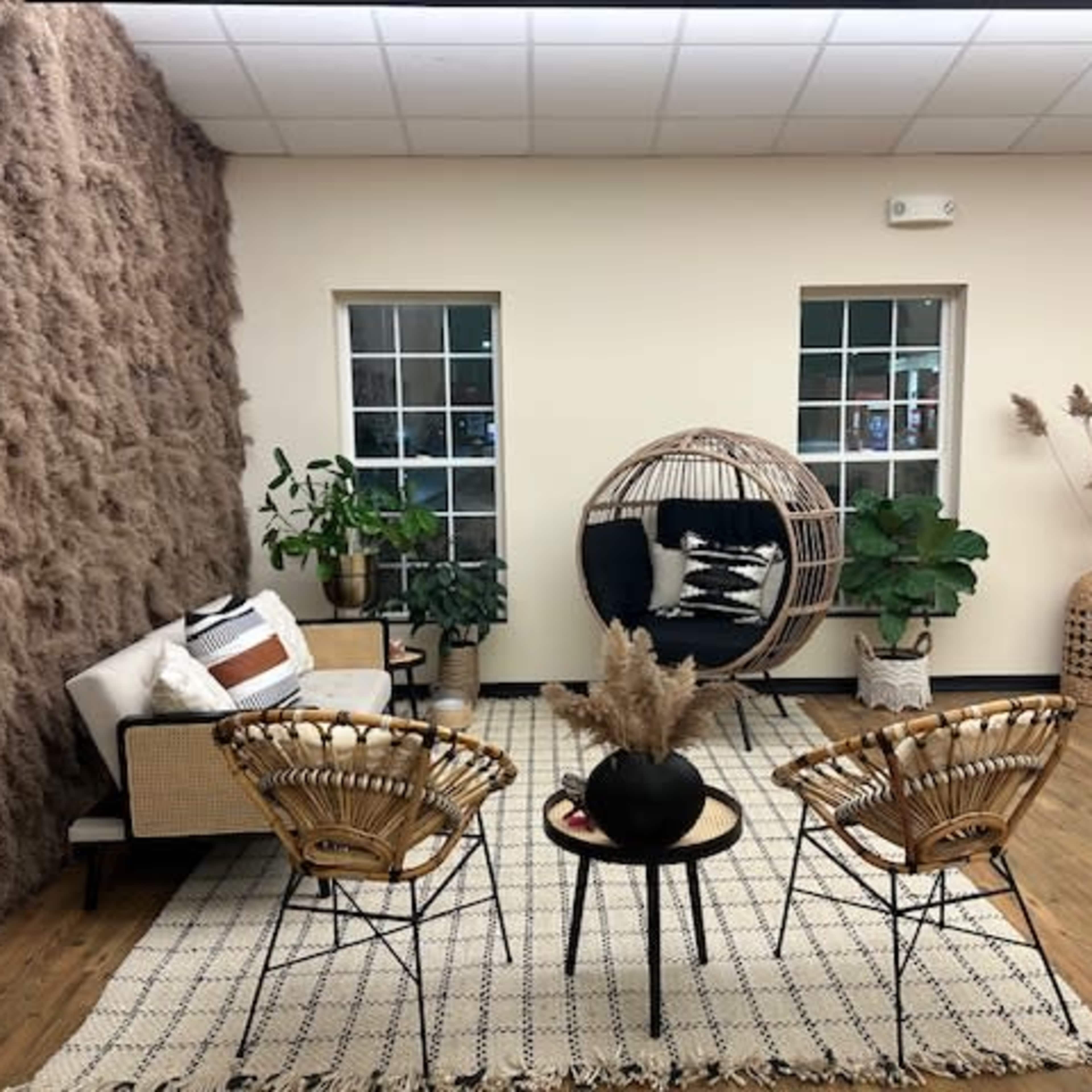 A cozy interior space featuring a hanging chair, a sofa, two woven chairs, and several plants arranged around a patterned rug on the wooden floor.
