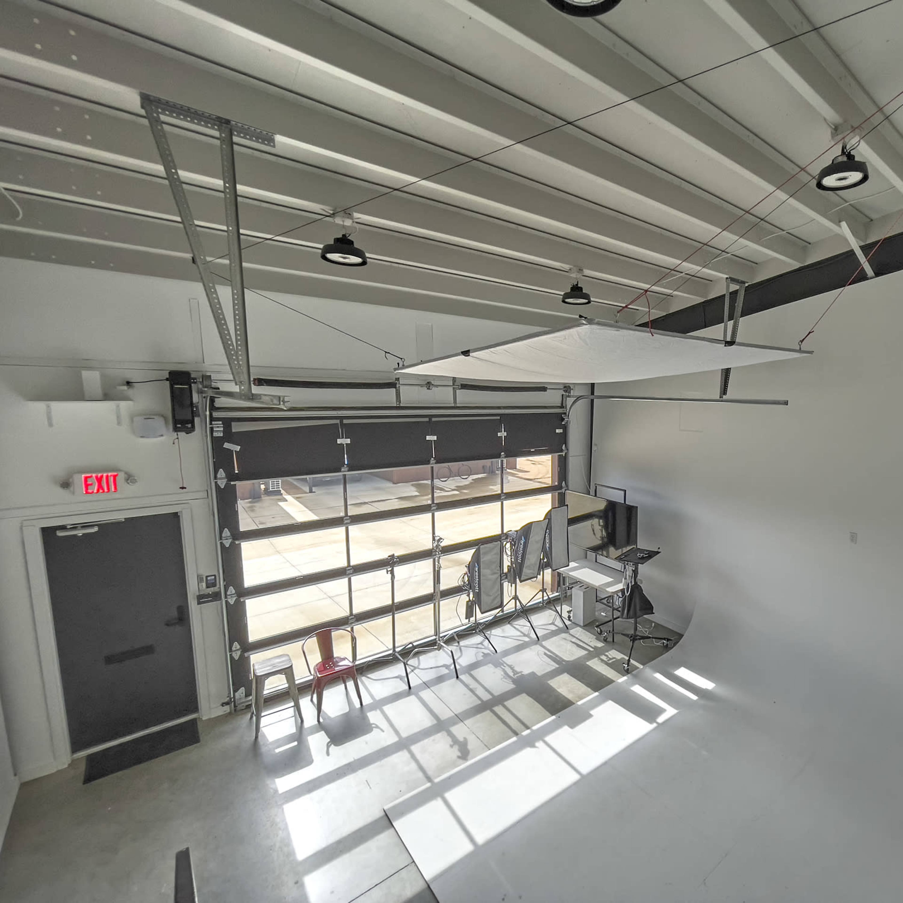 An empty studio with a large white sheet suspended from the ceiling, sunlight casting shadows on the floor through an open garage door.