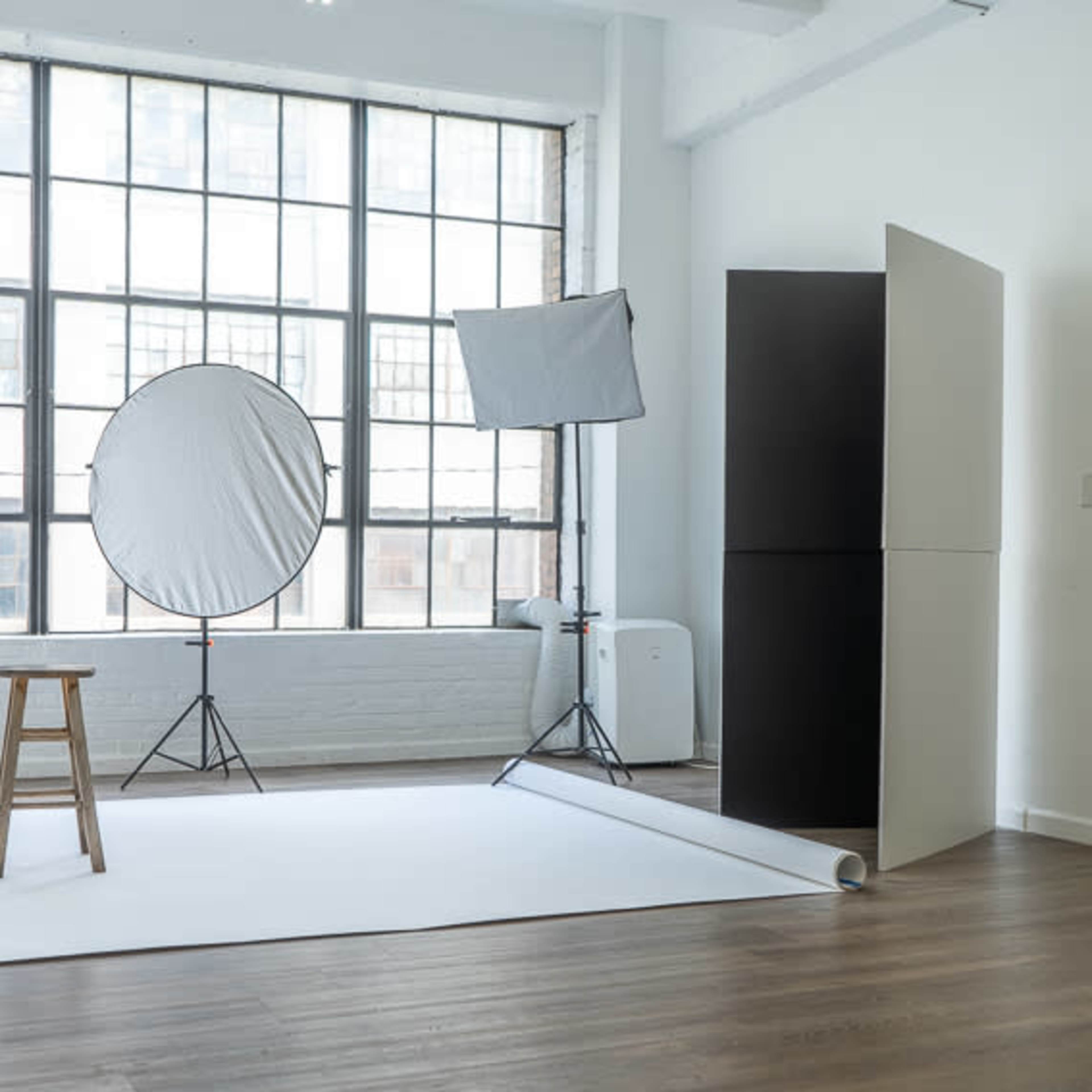 A photography studio with a white backdrop, a stool, lighting equipment, and large windows allowing natural light to enter.