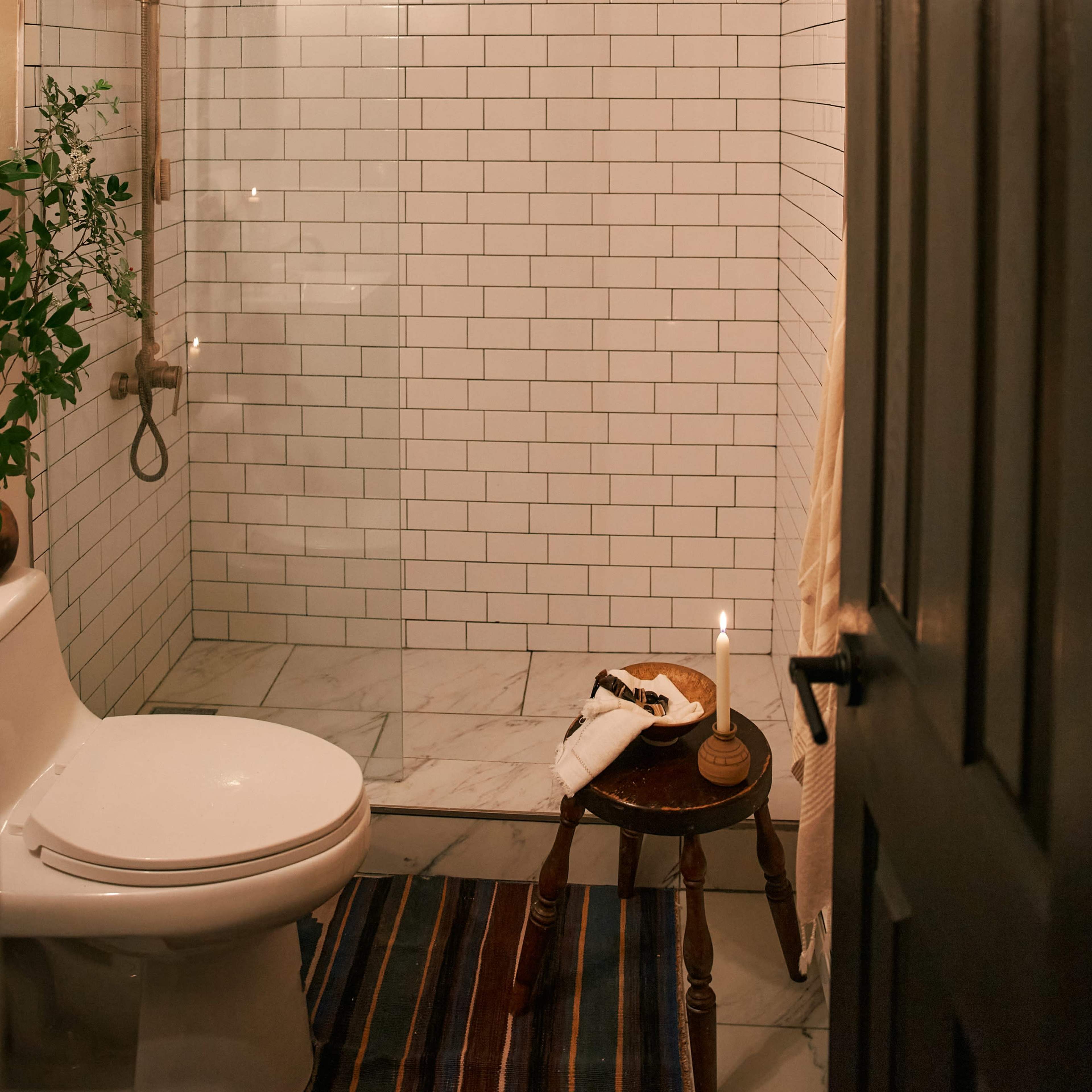 A bathroom with a glass shower enclosure, subway tile walls, a toilet, and a small table with a candle and a plate, all set on a marble floor.