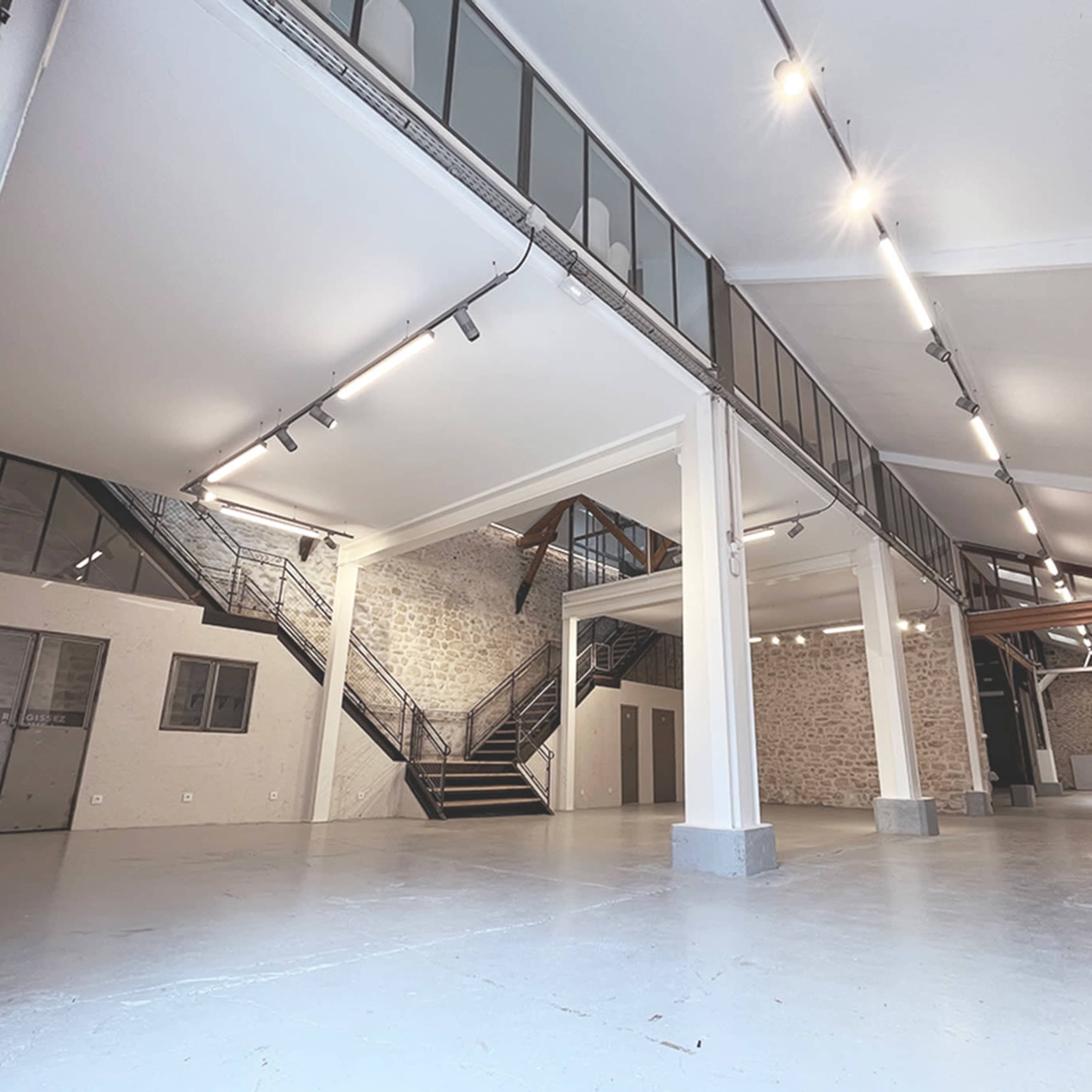 An open, well-lit industrial space with high ceilings, featuring exposed brick walls, a staircase, and minimalist design elements.