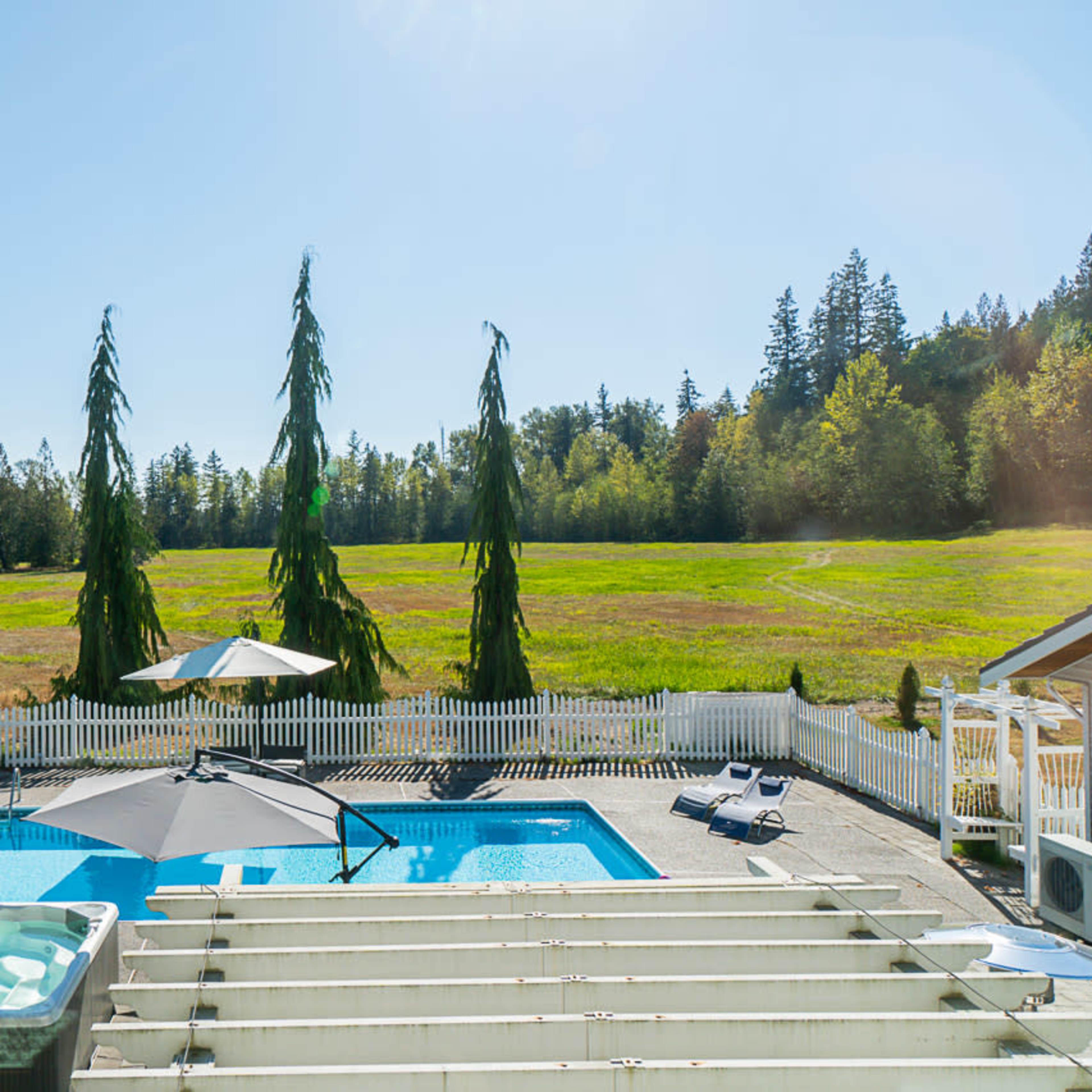 A backyard with a swimming pool, a hot tub, and a patio umbrella, surrounded by a white fence and tall evergreen trees, with a grassy field in the background.