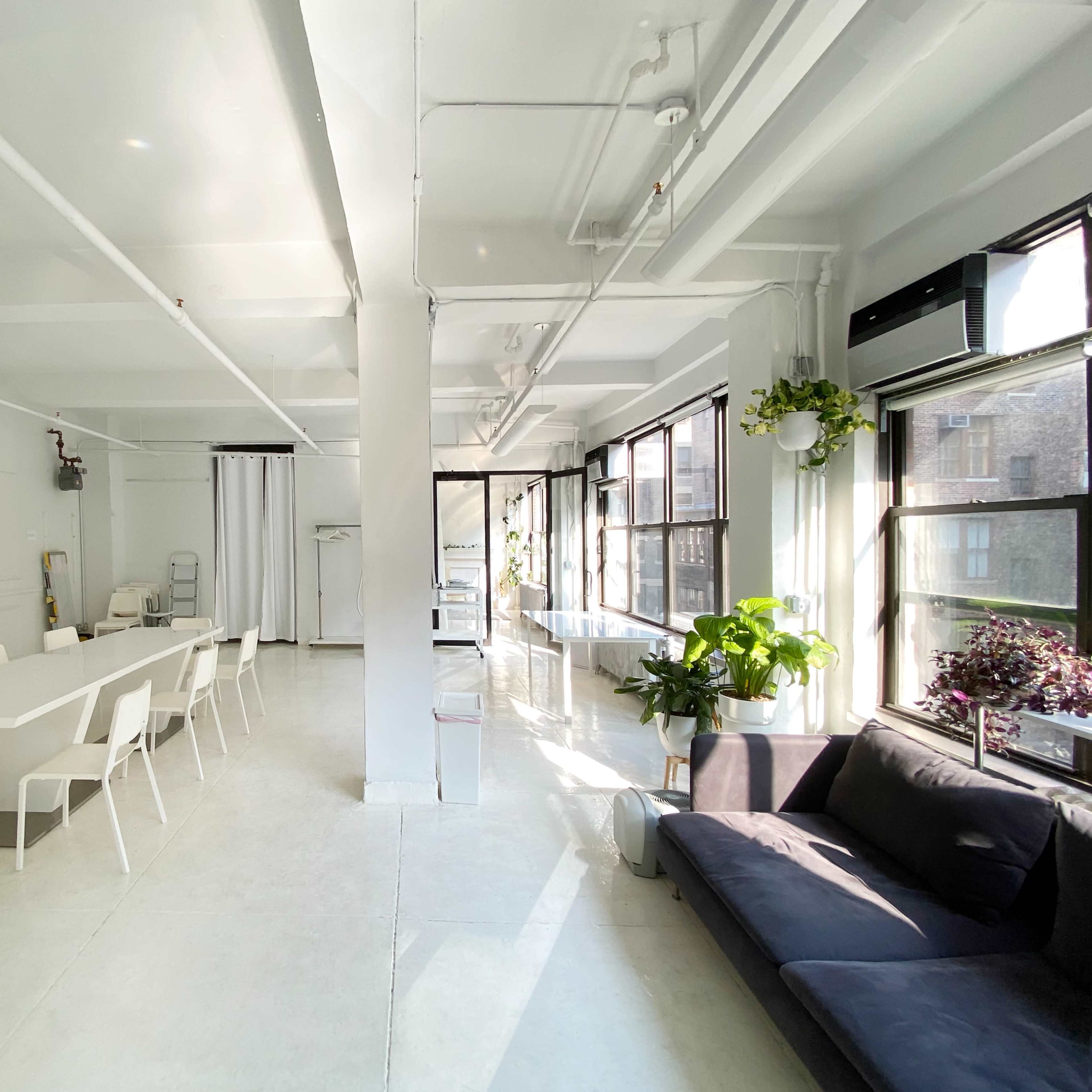 A bright, minimalist interior features a long white table surrounded by chairs, with large windows and greenery enhancing the space.