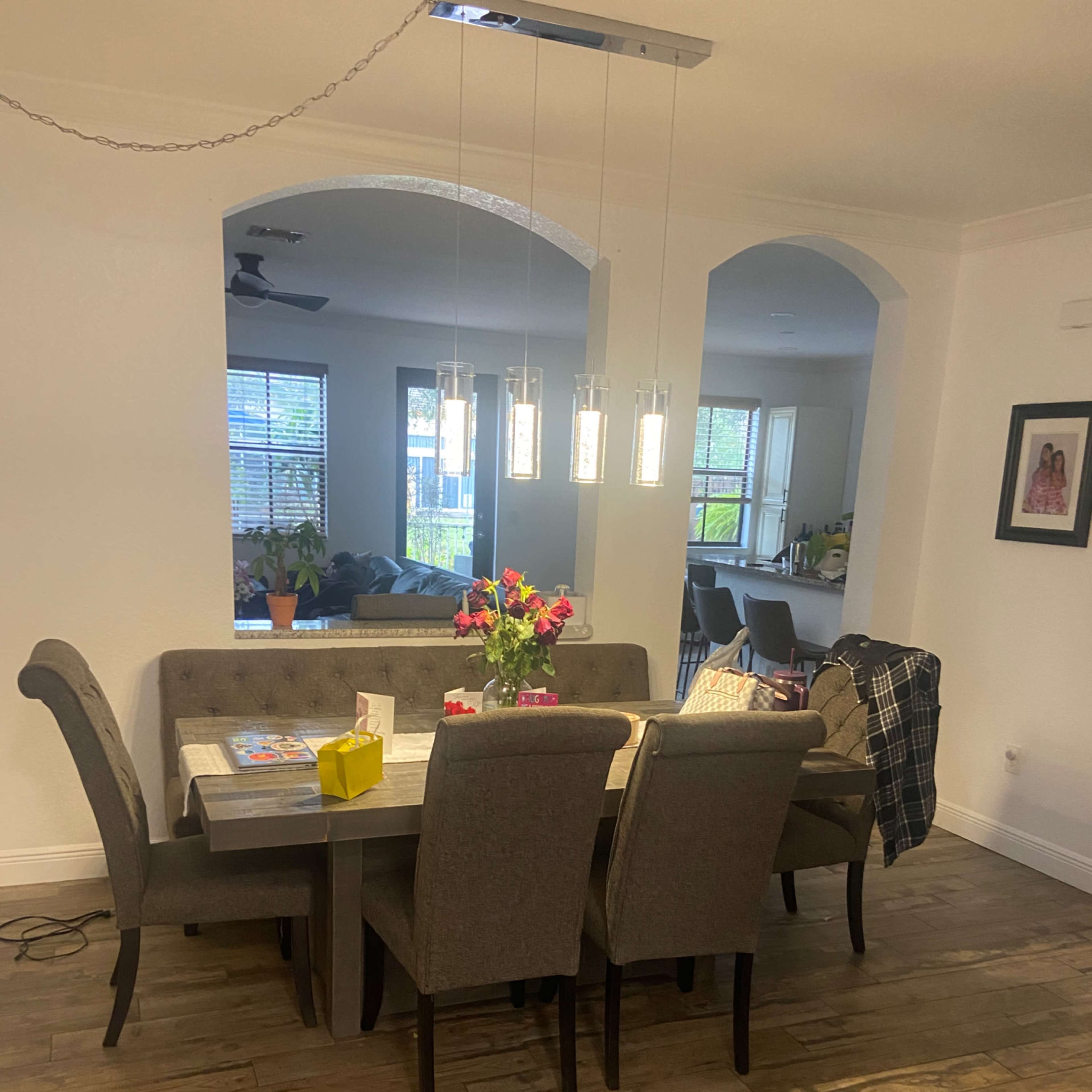 A dining area features a rectangular table with gray chairs, a vase of flowers, and modern pendant lights hanging above it, adjacent to a spacious living area with a sofa and large windows.
