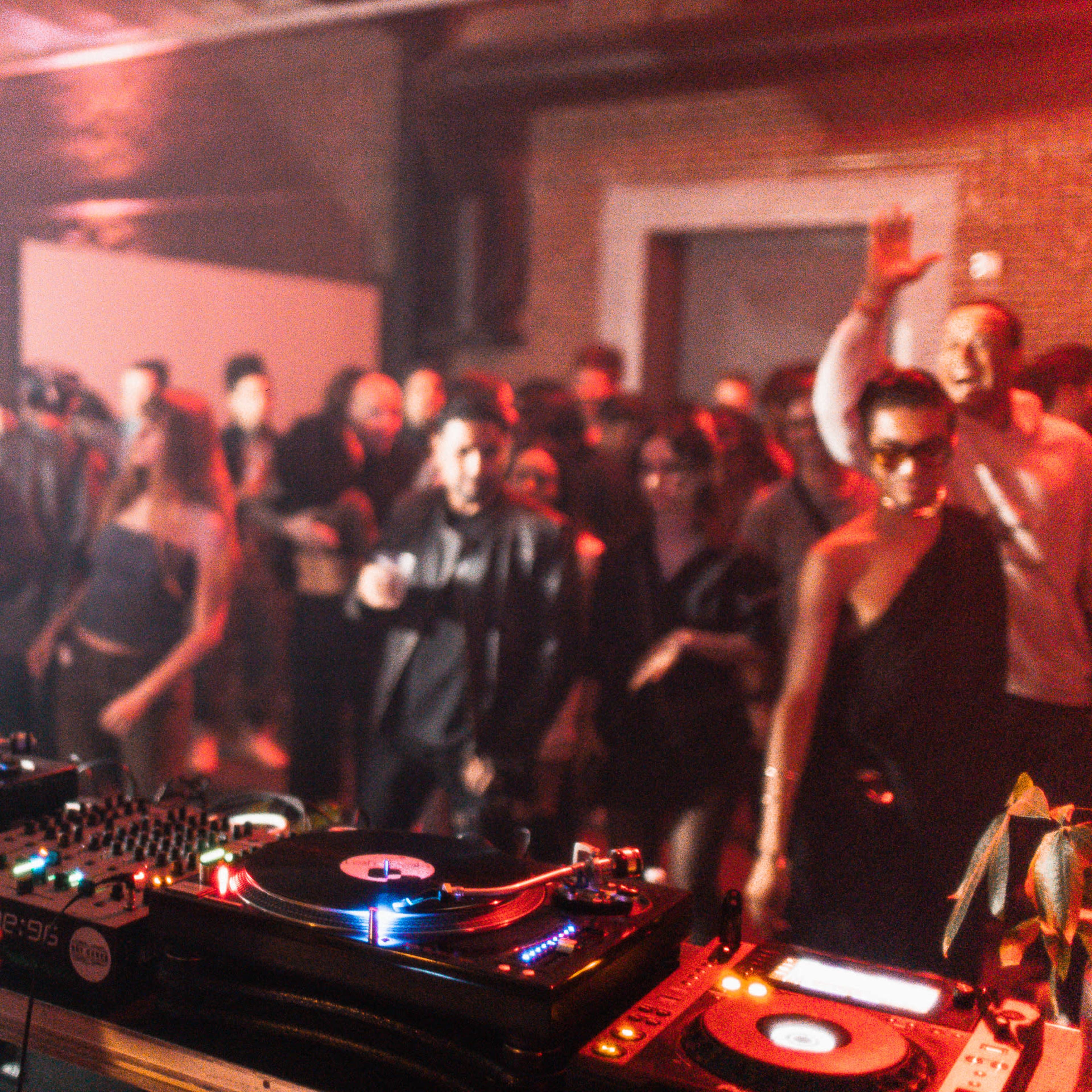 A DJ stands behind turntables in a dimly lit venue while an engaged crowd dances and cheers in the background.