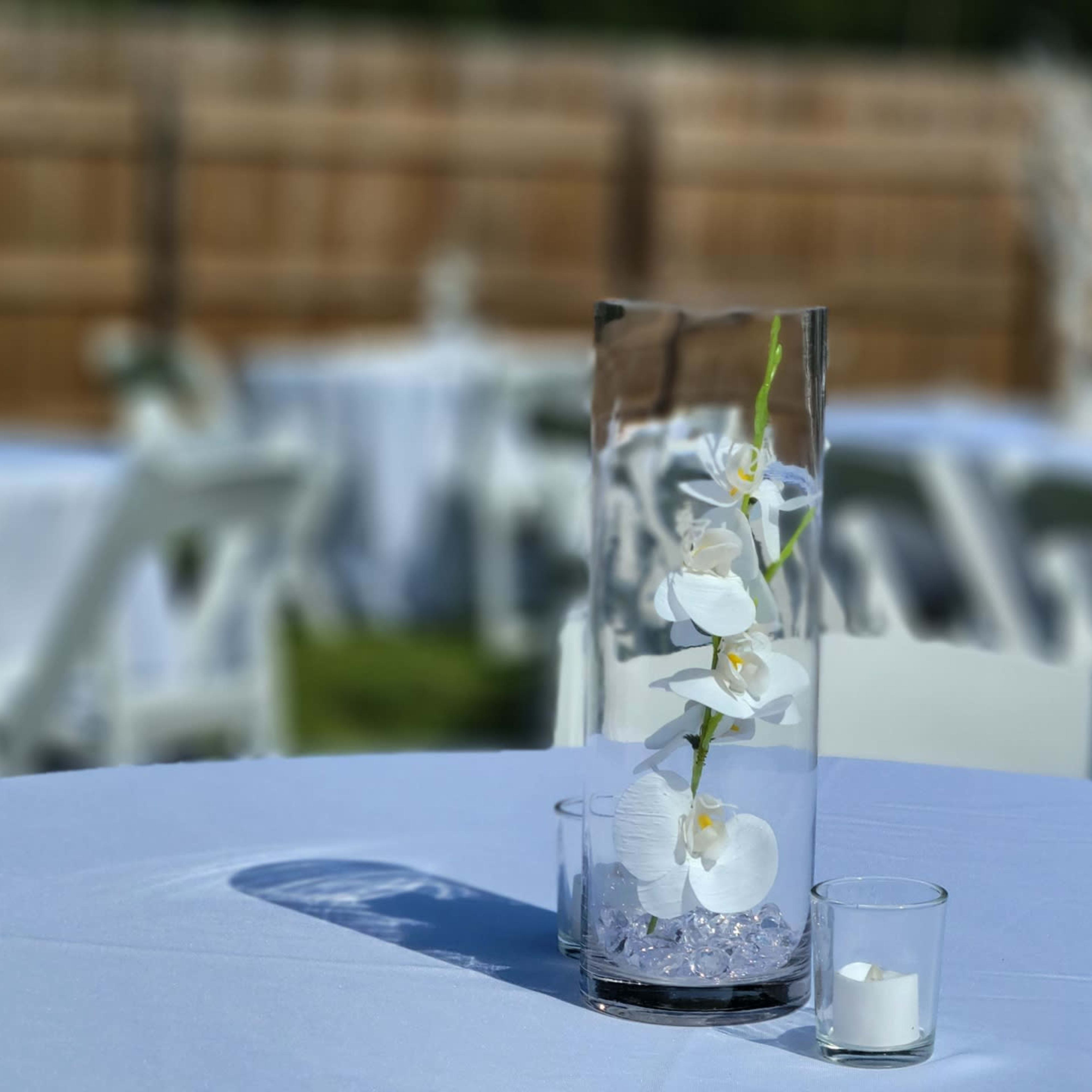 A clear vase with white orchids and small stones is placed on a table set for an event, surrounded by empty chairs in the background.
