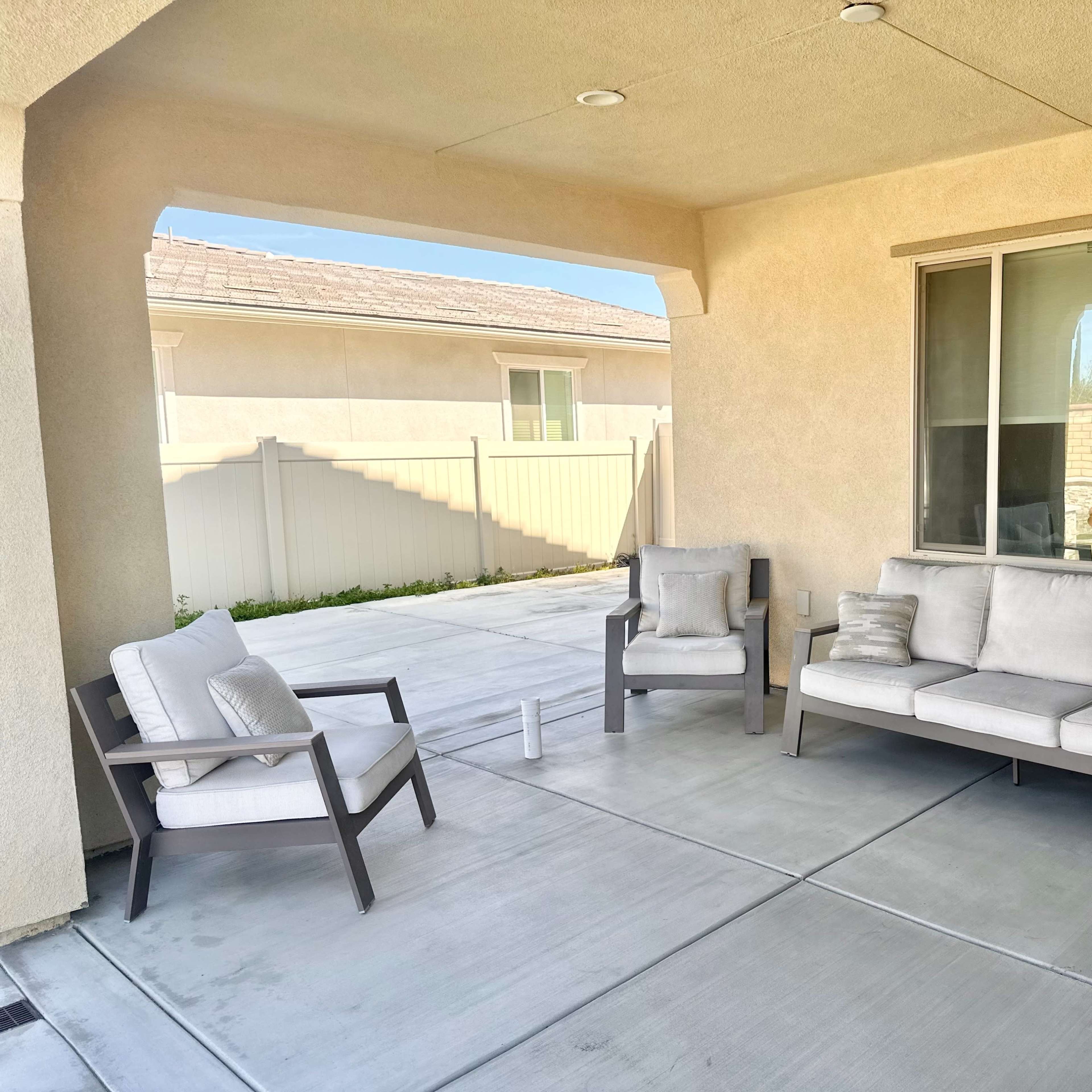 A covered patio area with two armchairs and a sofa arranged on a concrete floor, with a view of a backyard fence and a house in the background.